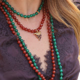 A woman wears two beaded necklaces, one red and the other green with a delicate grape charm. The necklaces are styled with a beautiful purple lace top for a bold and elegant look.