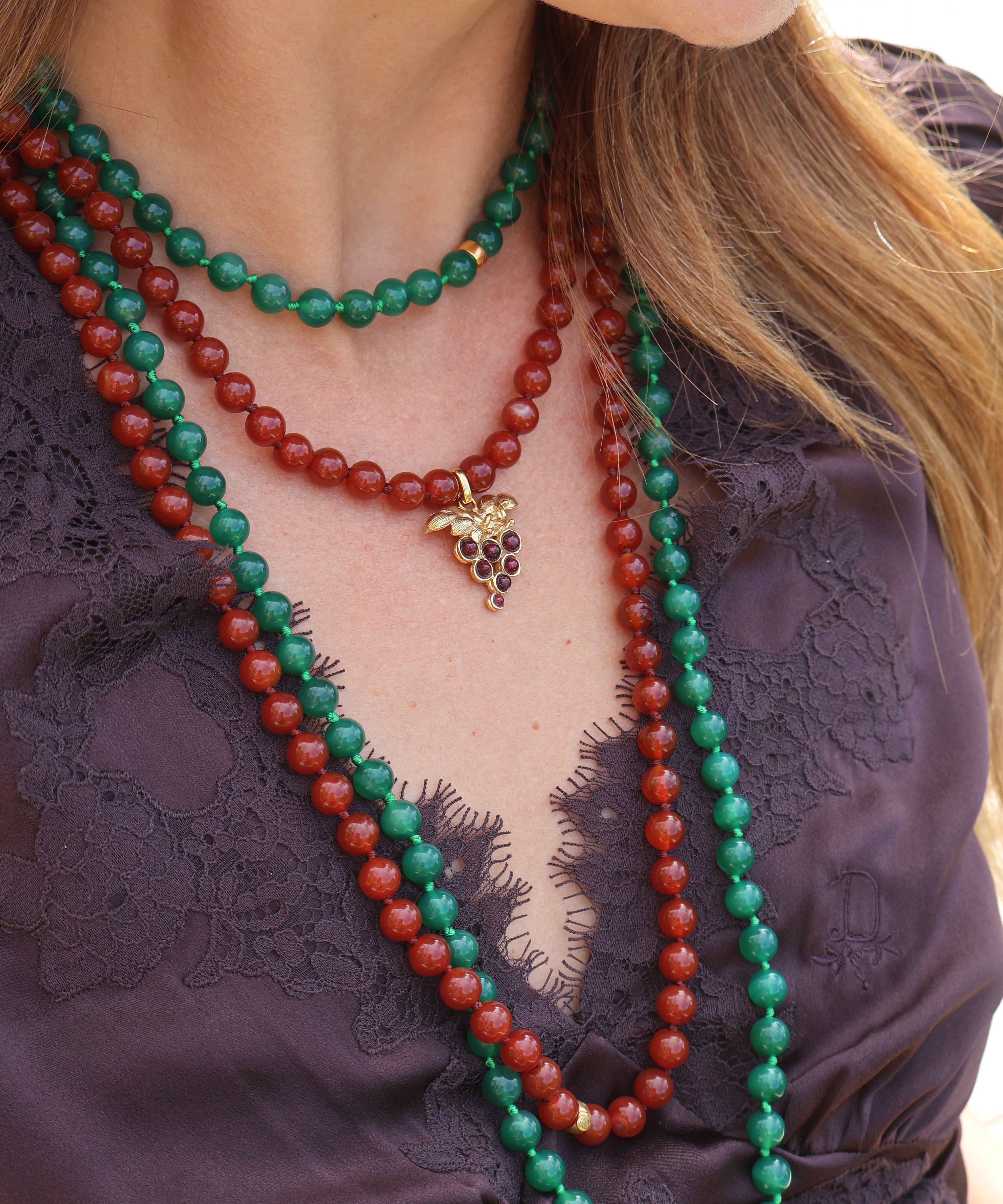 A woman wears two beaded necklaces, one red and the other green with a delicate grape charm. The necklaces are styled with a beautiful purple lace top for a bold and elegant look.