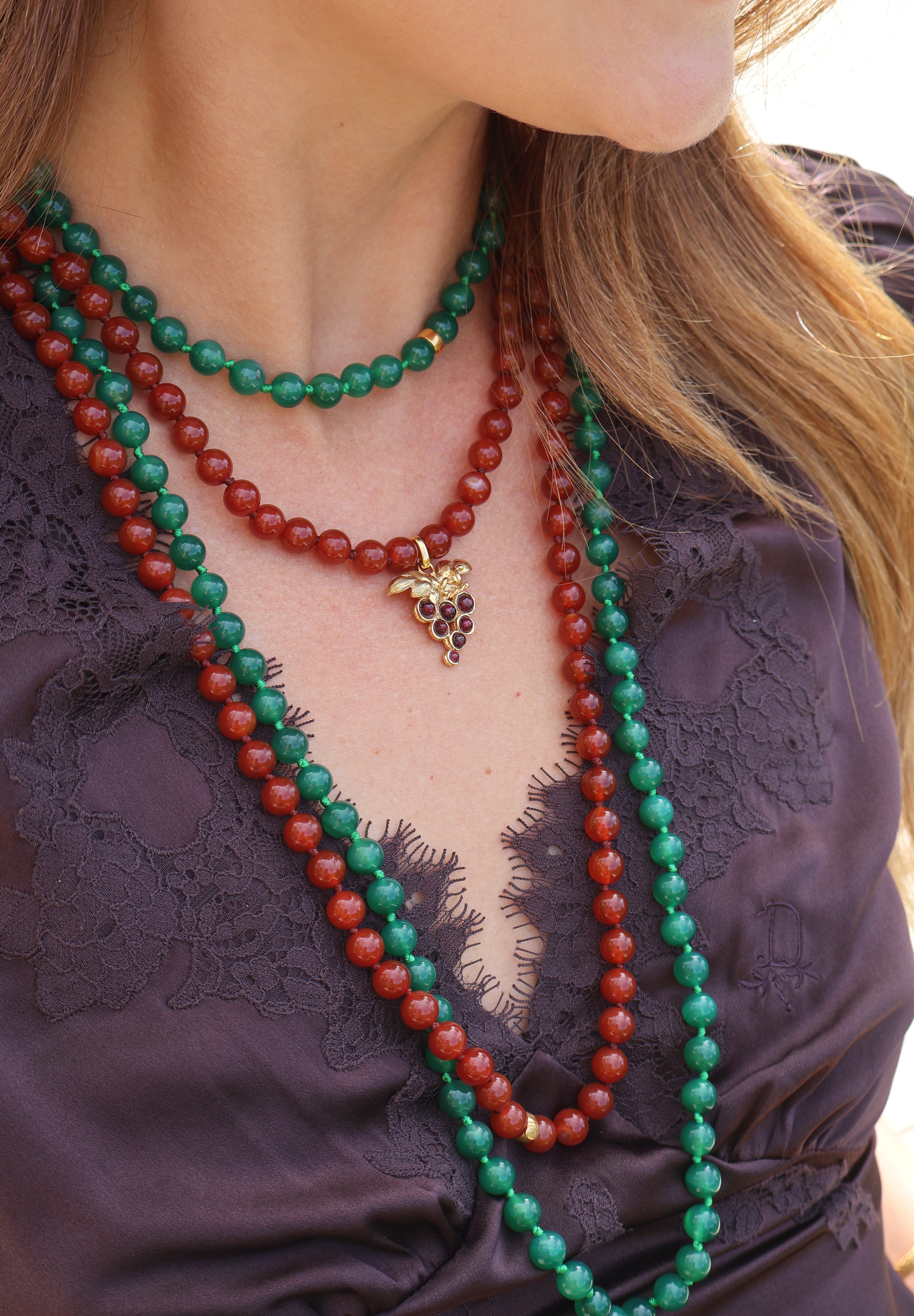 A woman wears two beaded necklaces, one red and the other green with a delicate grape charm. The necklaces are styled with a beautiful purple lace top for a bold and elegant look.