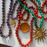 A close-up of carnelian, agate and amethyst bead necklaces  arranged on a light fabric surface. The necklaces feature purple, red, and green beads with gold-toned pendants, including a round amber pendant and a sunburst pendant.
