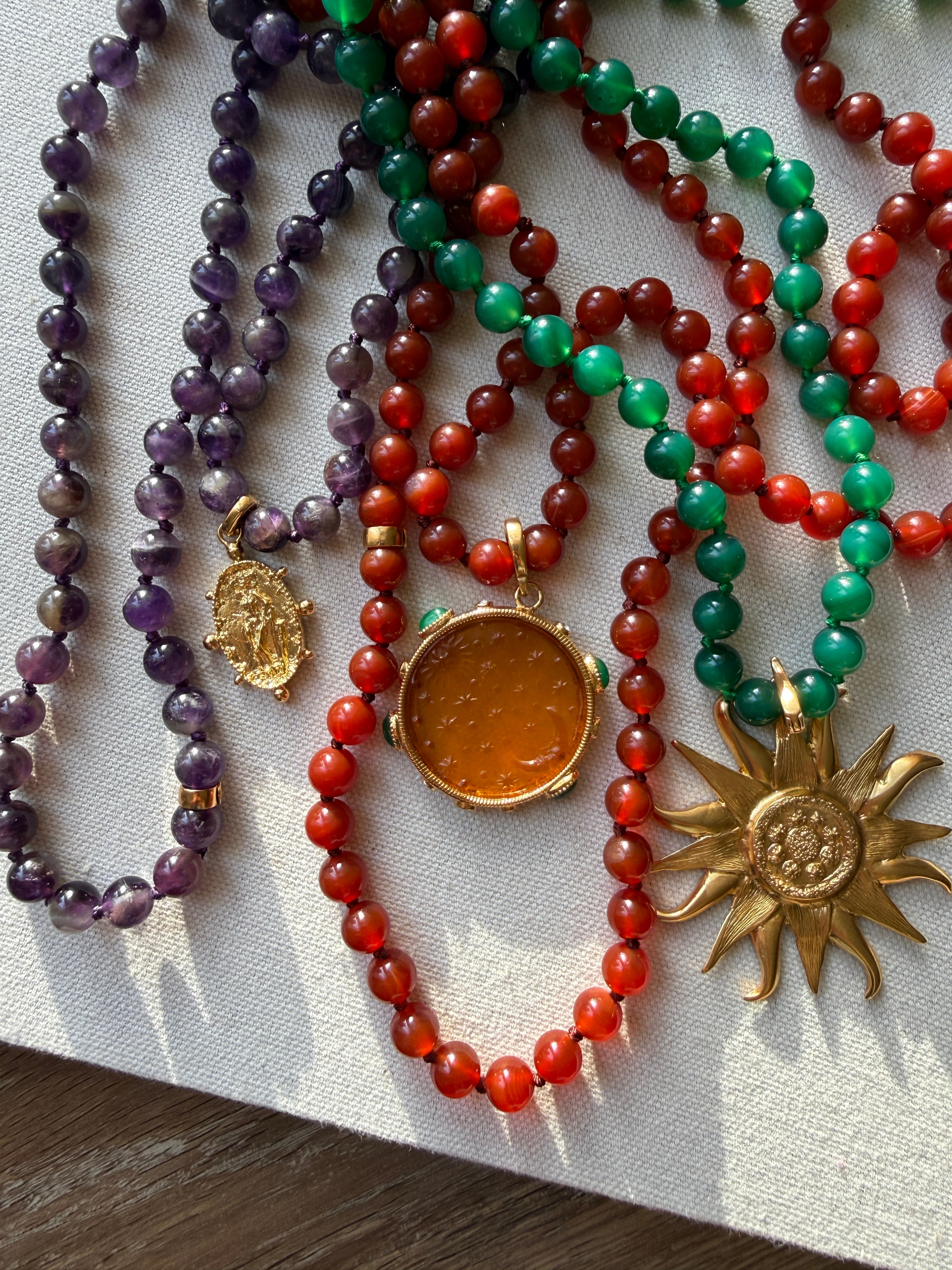 A close-up of carnelian, agate and amethyst bead necklaces  arranged on a light fabric surface. The necklaces feature purple, red, and green beads with gold-toned pendants, including a round amber pendant and a sunburst pendant.
