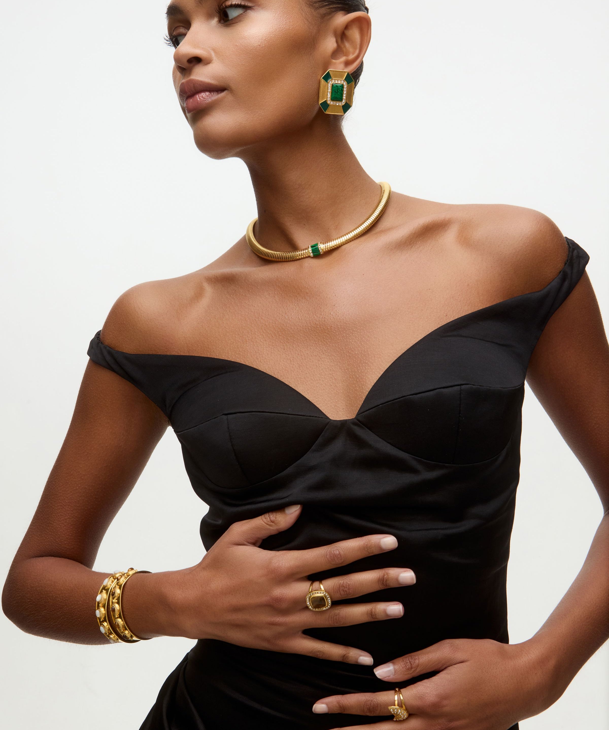 Woman wearing a black dress with matching gold and green earrings and necklace on a neutral background