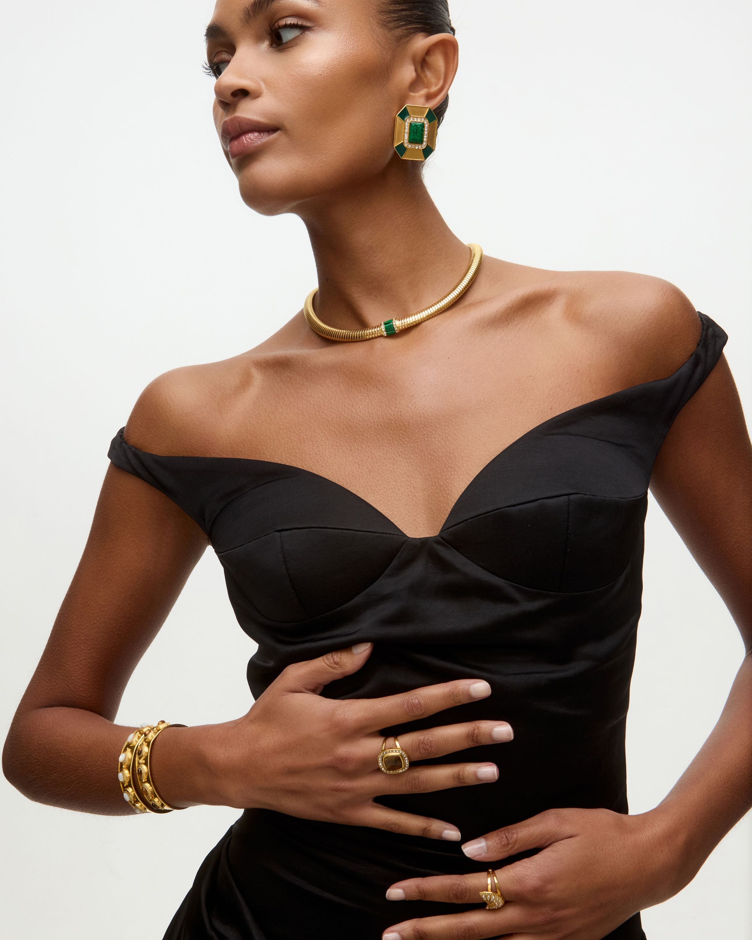 Woman wearing a black dress with matching gold and green earrings and necklace on a neutral background