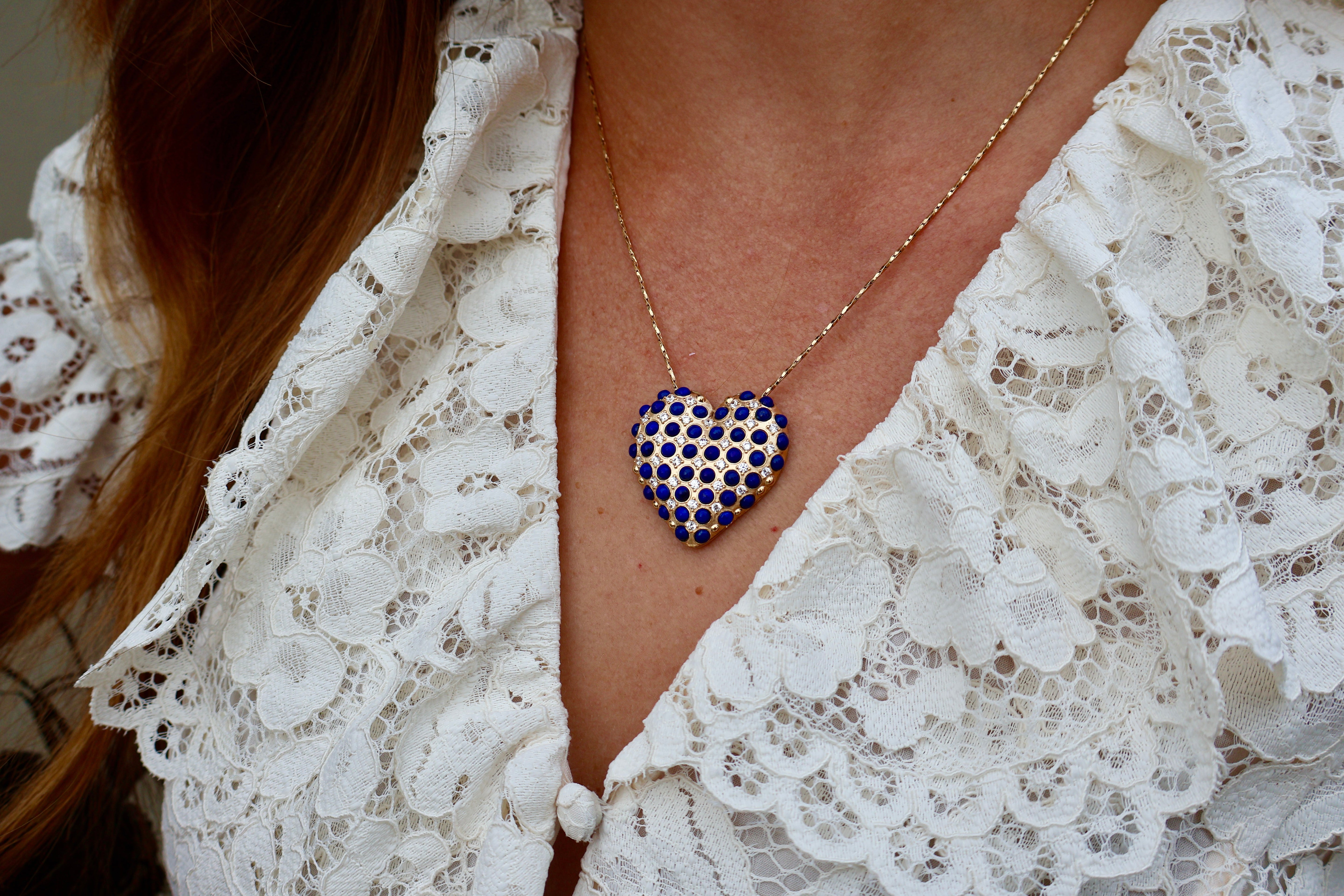 A close-up lifestyle shot of a person wearing a heart-shaped pendant necklace embellished with blue lapis beads and sparkling clear crystals, suspended from a delicate gold chain and styled with an elegant white lace blouse.
