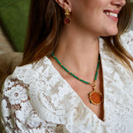 A woman wearing a gold and green beaded necklace with a round amber pendant, paired with amber drop earrings. She is smiling and shown in a close-up profile, with a cozy indoor background featuring a dog on a couch.