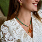 A woman wearing a gold and green beaded necklace with a round amber pendant, paired with amber drop earrings. She is smiling and shown in a close-up profile, with a cozy indoor background featuring a dog on a couch.