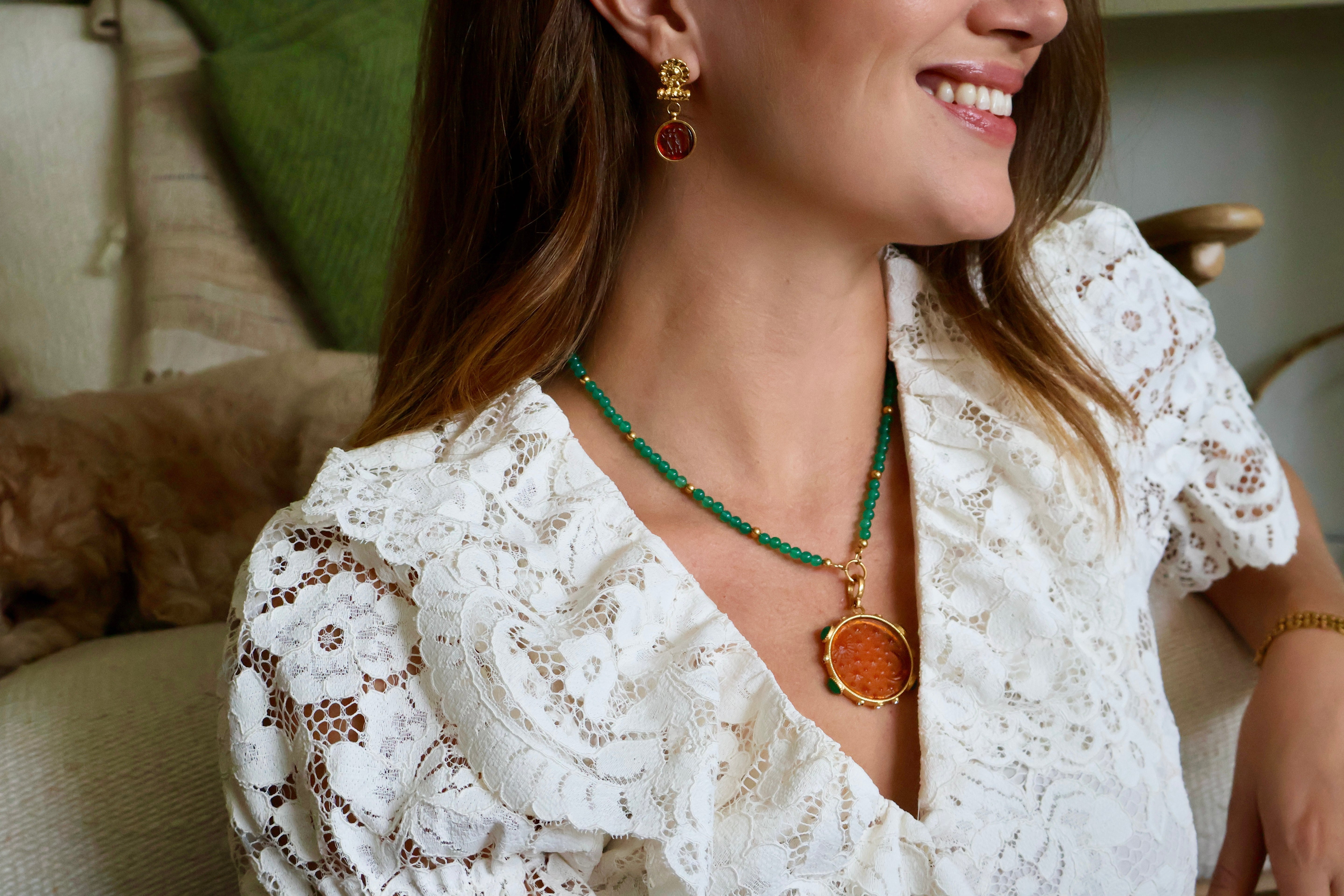 A woman wearing a gold and green beaded necklace with a round amber pendant, paired with amber drop earrings. She is smiling and shown in a close-up profile, with a cozy indoor background featuring a dog on a couch.
