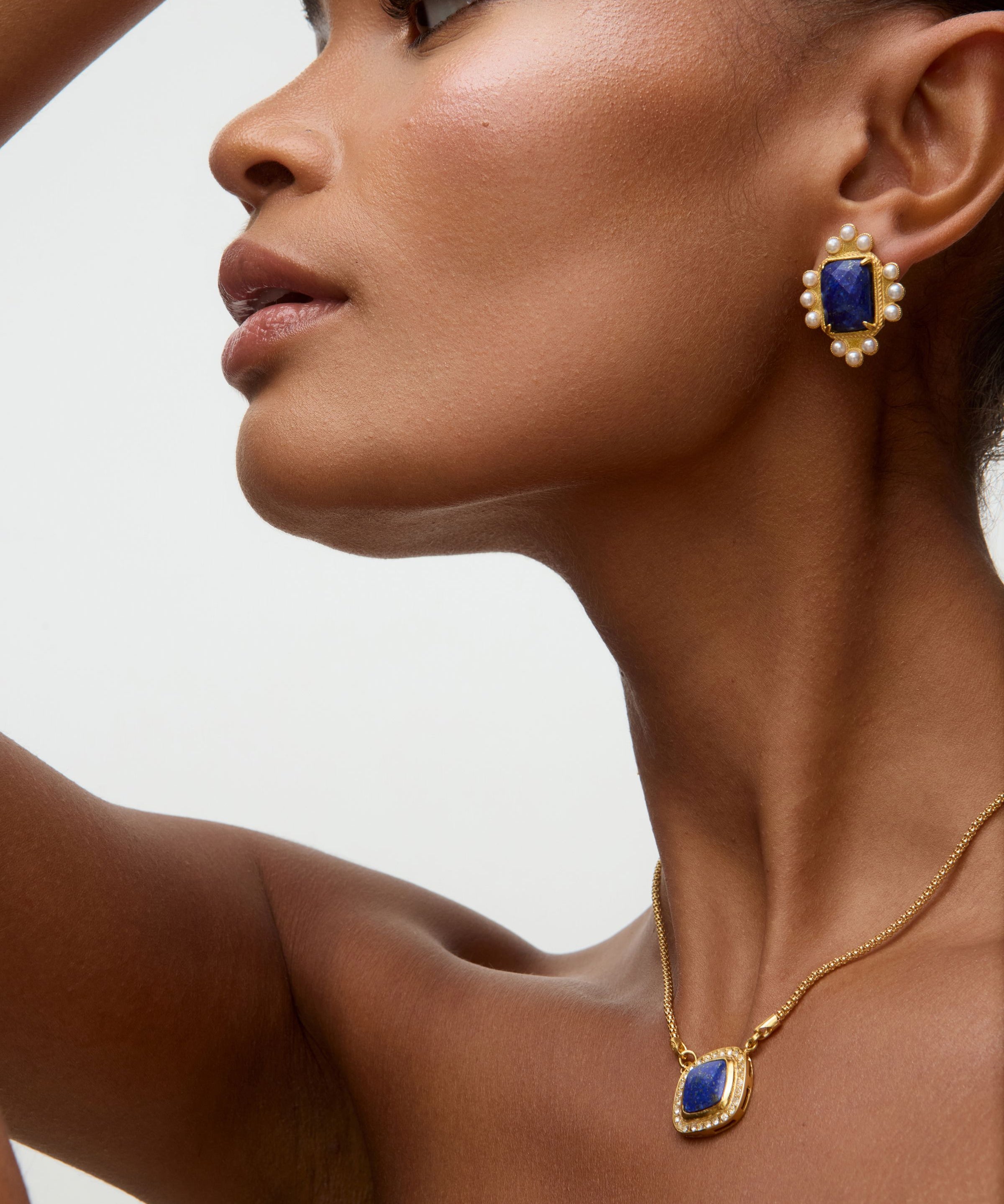 Close-up of a woman wearing gold earrings and necklace with blue stones on a neutral background