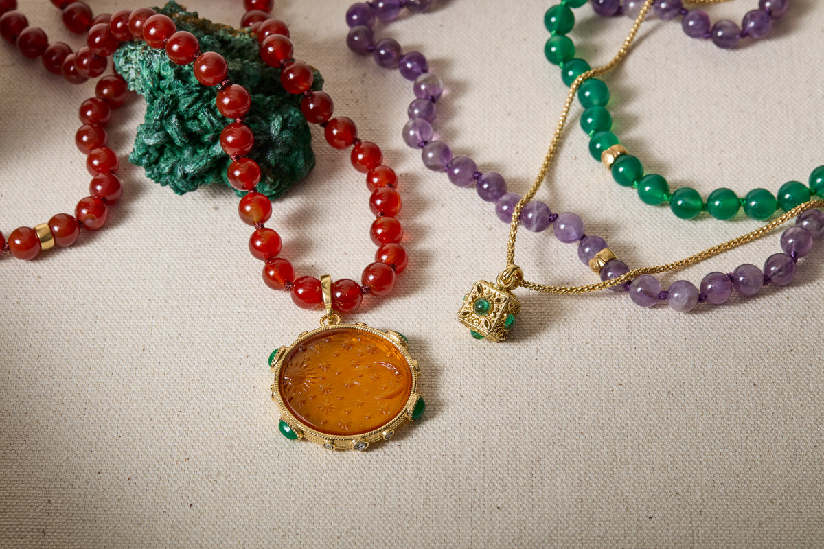A close-up flat lay of several beaded necklaces on a light fabric background. One necklace has translucent red beads with a round amber-orange pendant in a gold frame.