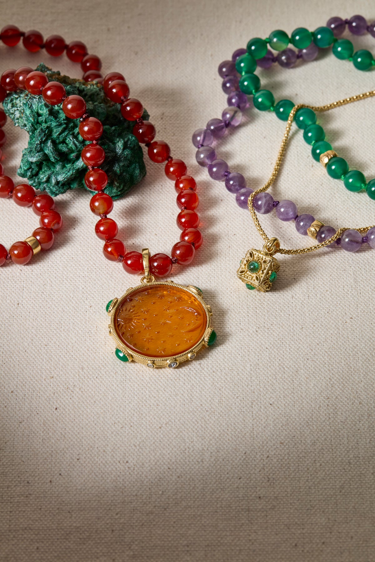 A close-up flat lay of several beaded necklaces on a light fabric background. One necklace has translucent red beads with a round amber-orange pendant in a gold frame.