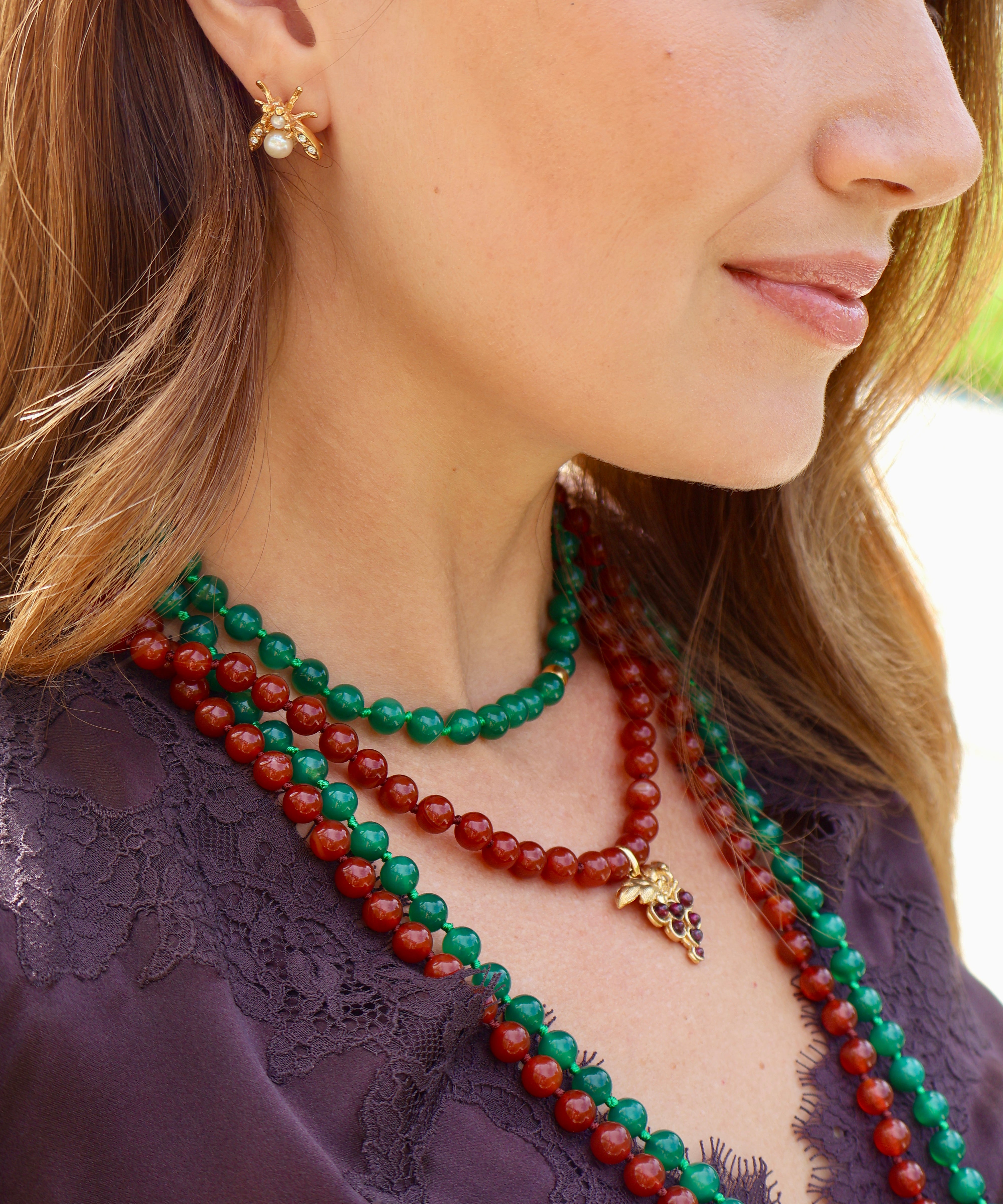 A close-up side profile of a woman wearing layered beaded necklaces and a matching gold earring. The necklaces include green and red beaded strands, with one necklace featuring a small gold grapevine pendant.