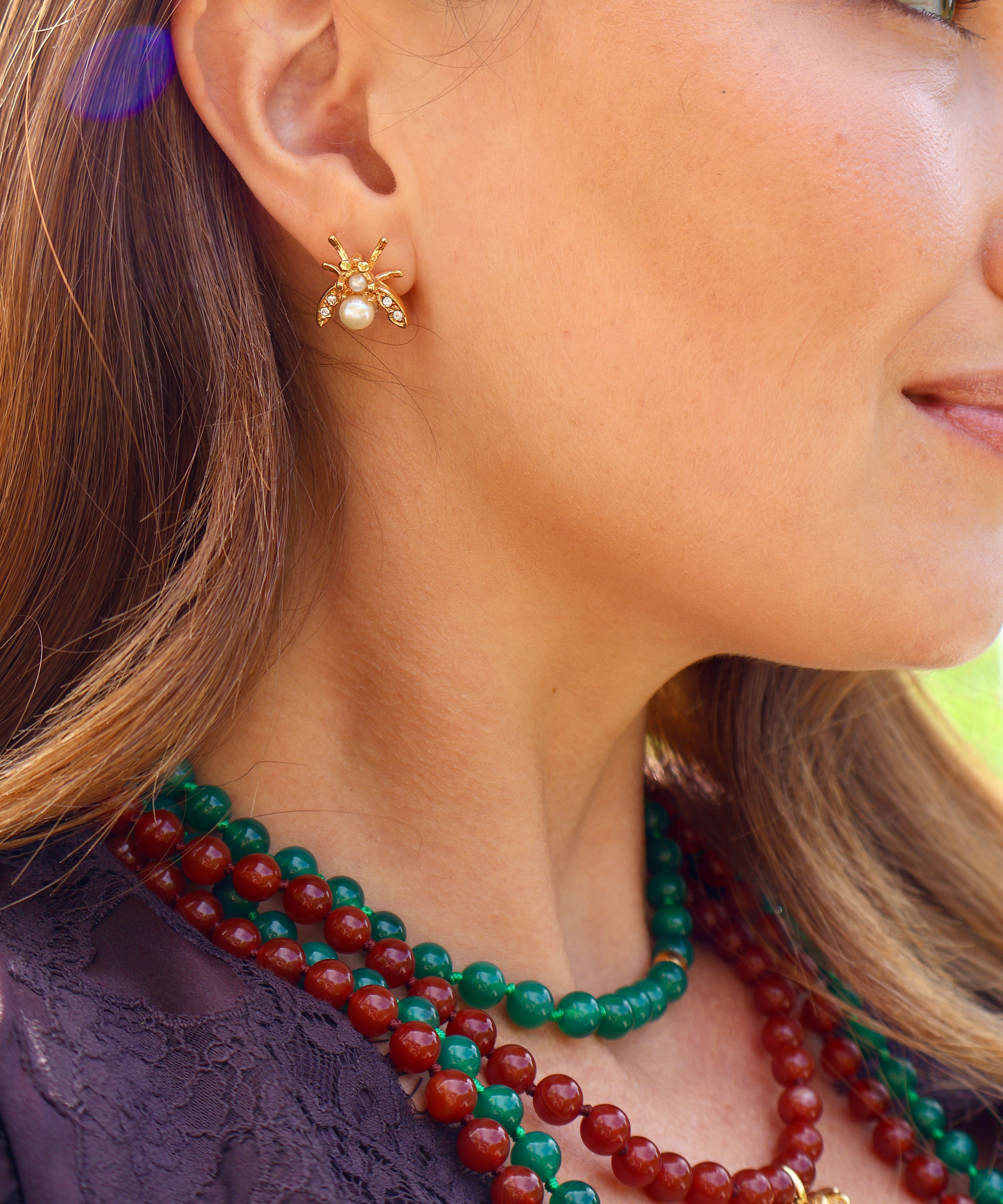 A close-up side profile of a woman wearing a small gold bee stud earring with a pearl center, layered with green and red beaded necklaces.