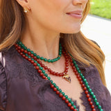 A woman wearing a small gold bee stud earring with a pearl centre, layered with green and red beaded necklaces.