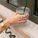Hand holding a drink with a straw, wearing gold jewelry, against a blurred indoor background.
