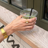 Hand holding a drink with a straw, wearing gold jewelry, against a blurred indoor background.