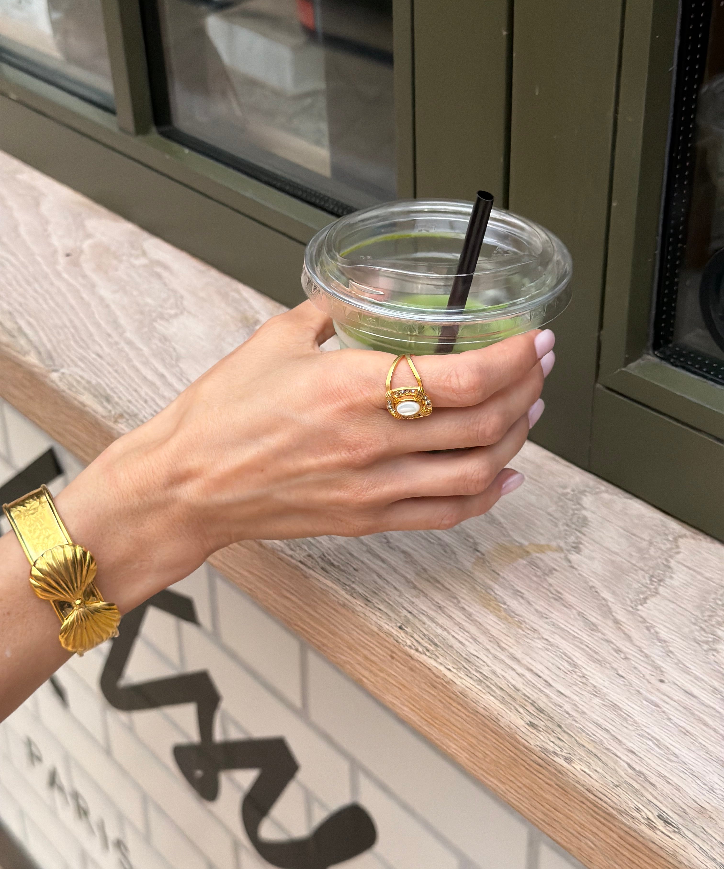 Hand holding a drink with a straw, wearing gold jewelry, against a blurred indoor background.