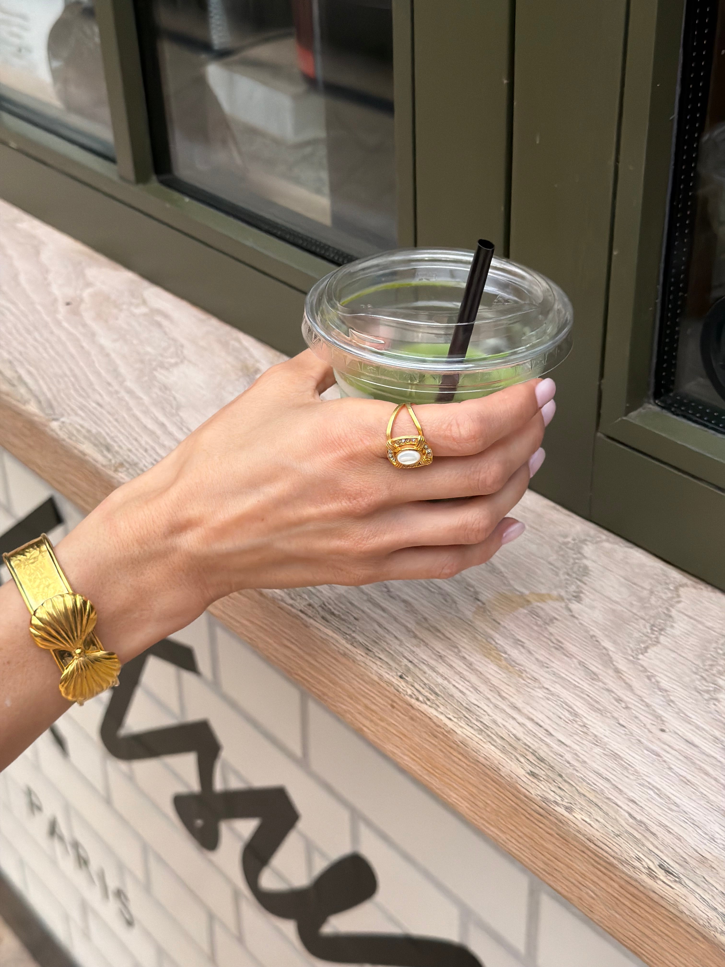 Hand holding a drink with a straw, wearing gold jewelry, against a blurred indoor background.
