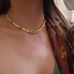 A close-up lifestyle shot of a woman wearing a gold chain necklace with a central green agate stone and small clear stones, styled with a green top and a rust-colored floral shirt.