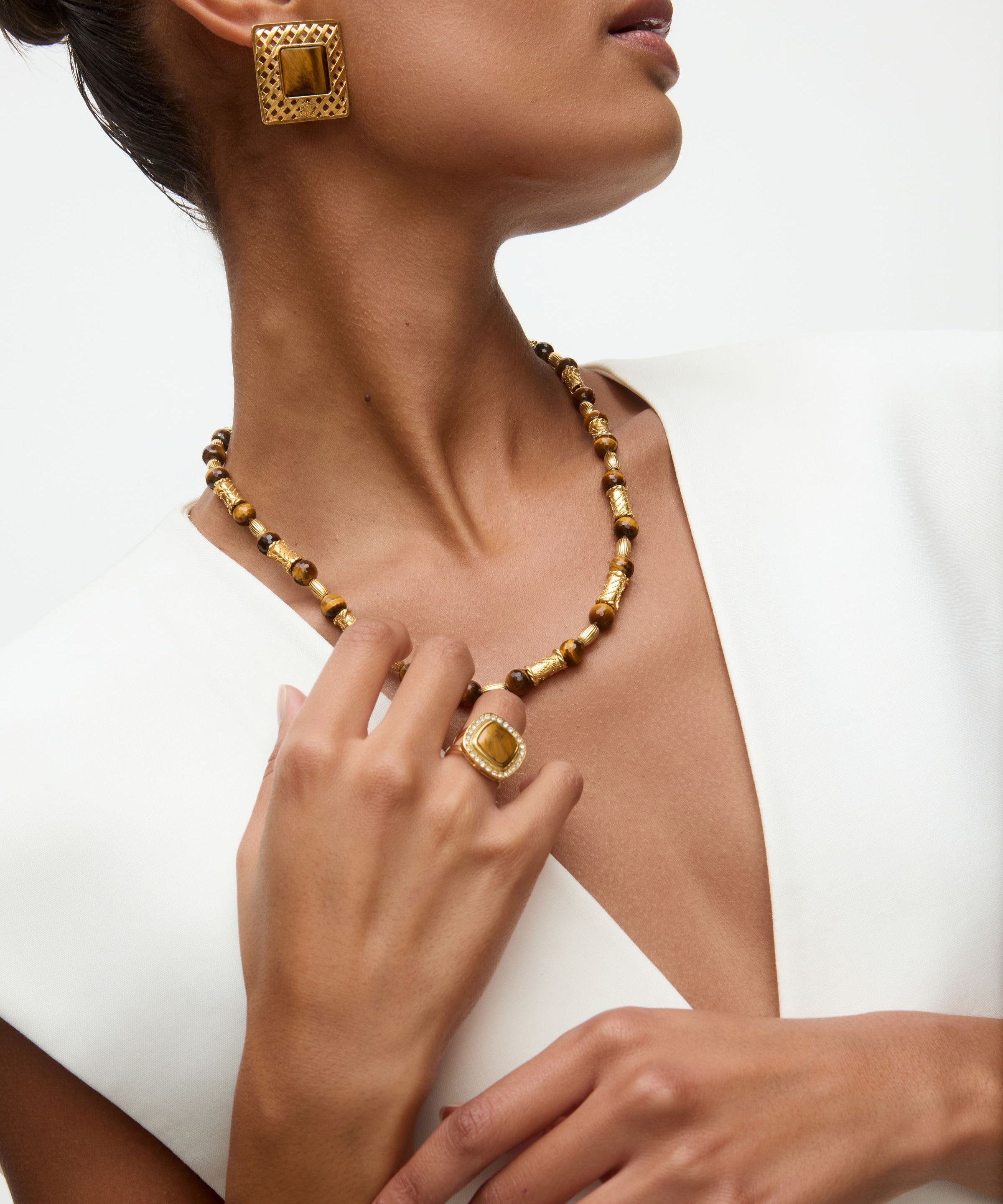 Close-up of a person wearing gold and tigers eye jewelry including a necklace, ring, and earrings on a white background.