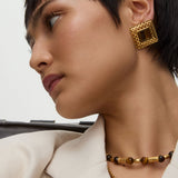 Close-up of a person wearing square gold earrings with tigers eye stone and a necklace on a neutral background