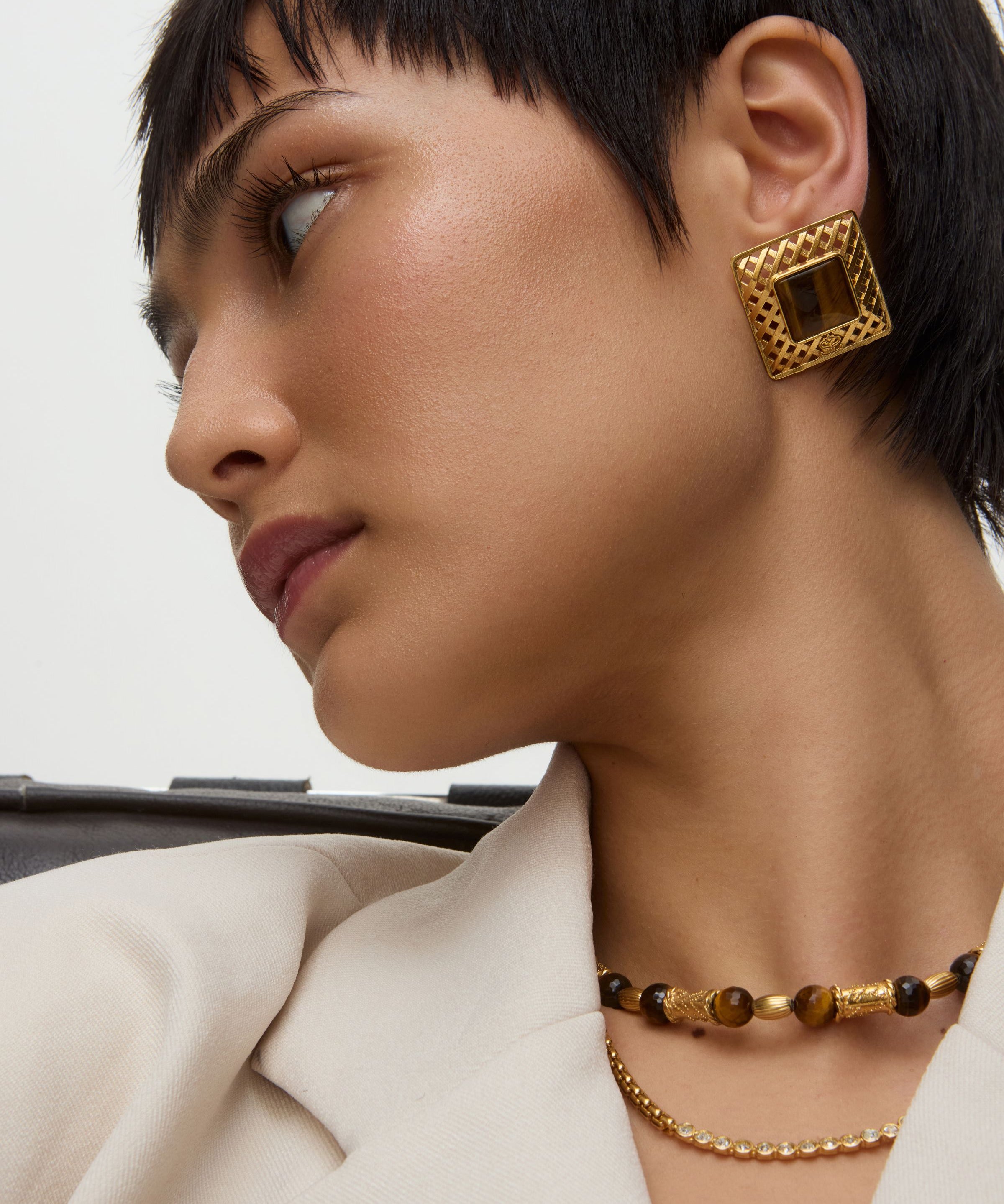 Close-up of a person wearing square gold earrings with tigers eye stone and a necklace on a neutral background