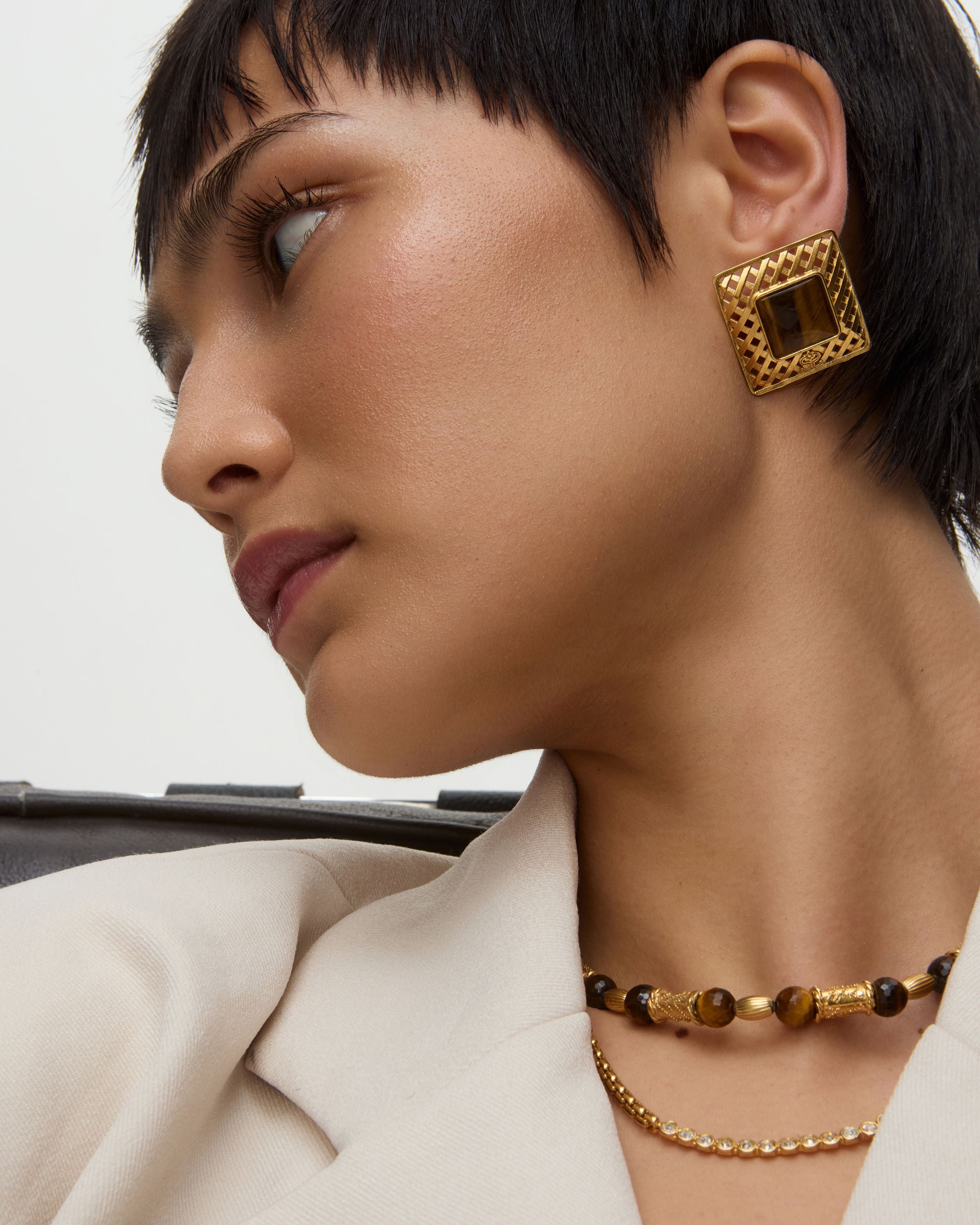Close-up of a person wearing square gold earrings with tigers eye stone and a necklace on a neutral background