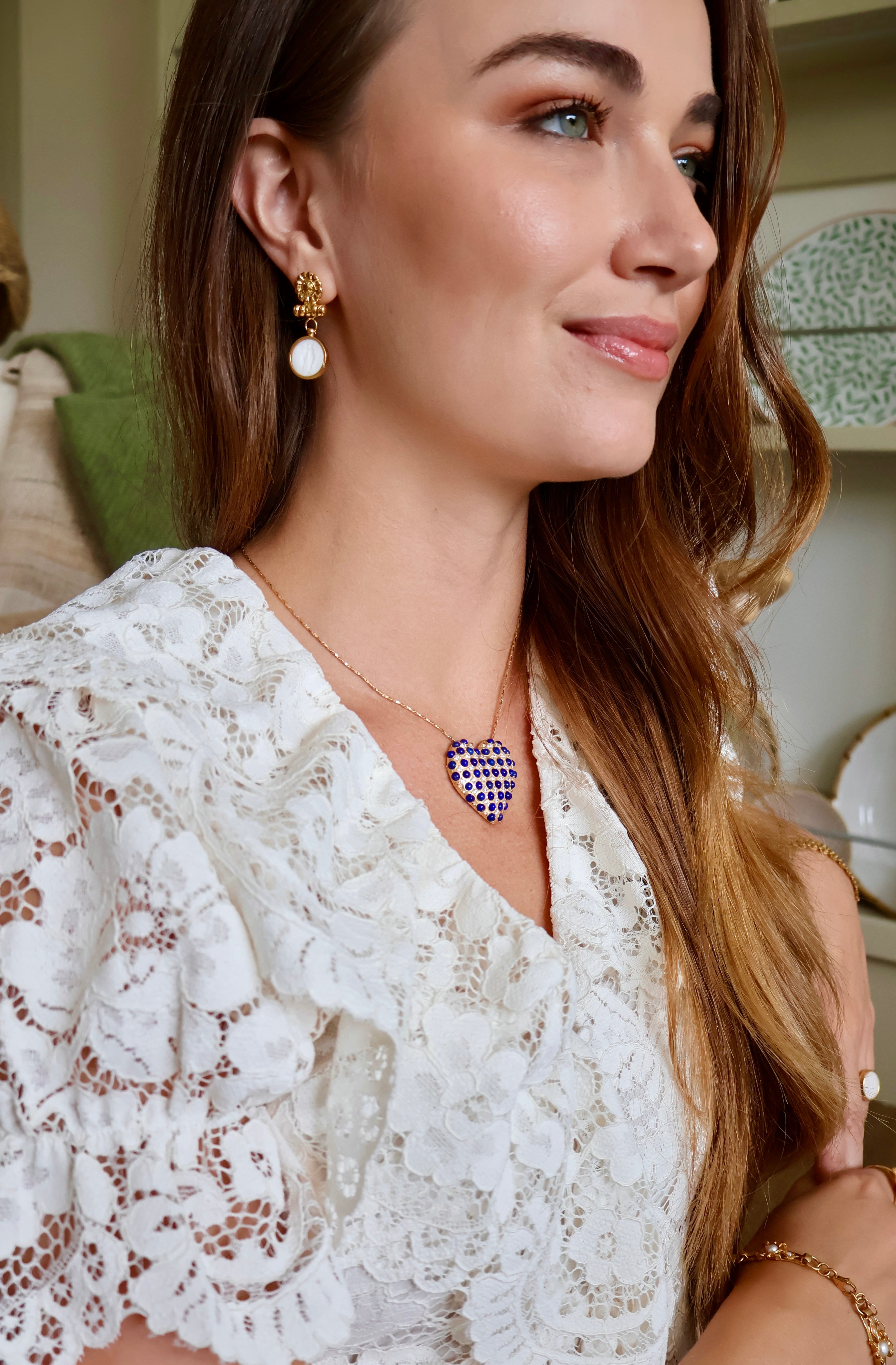 A woman wearing a gold necklace with a blue and white gingham heart pendant, styled with a white lace top in an indoor setting.