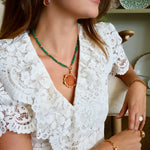 A woman wearing a gold and green necklace with a round amber pendant, a gold bracelet, and a matching gold earring, paired with a white lace top.