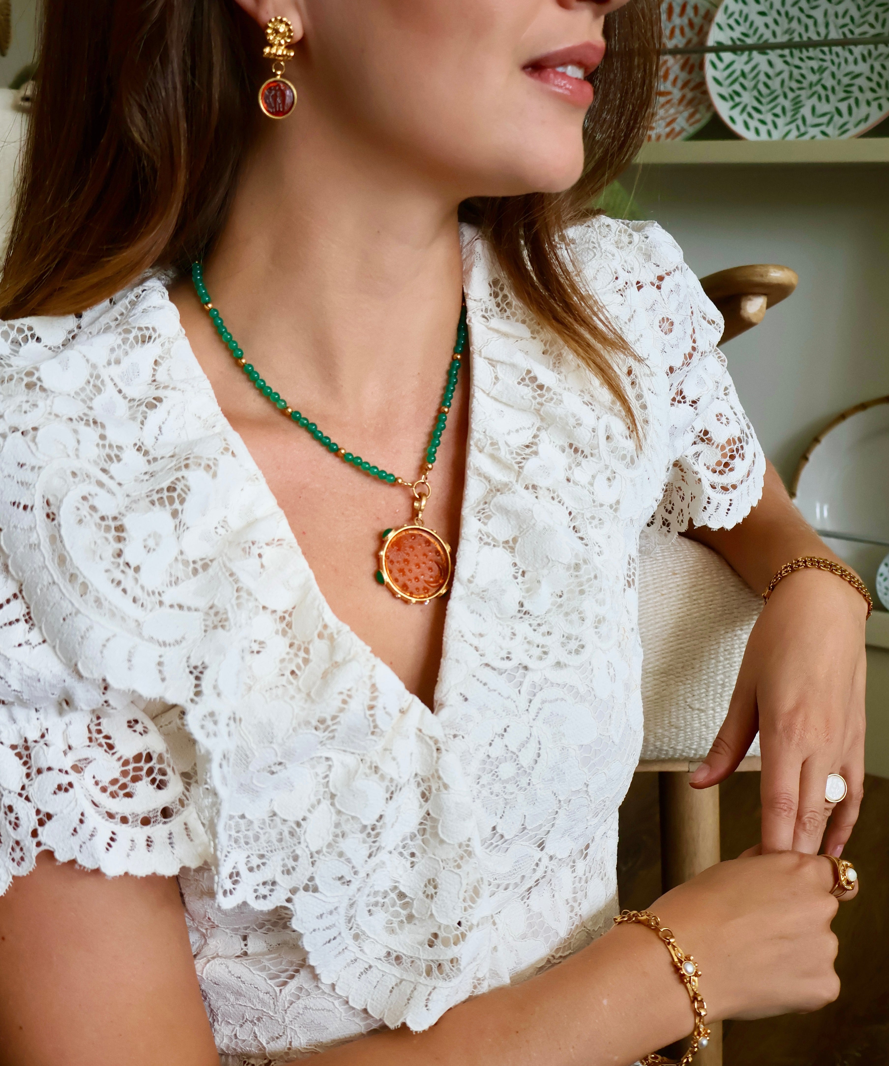 A woman wearing a gold and green necklace with a round amber pendant, a gold bracelet, and a matching gold earring, paired with a white lace top.
