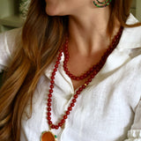 A close-up lifestyle shot of a woman wearing a long red beaded necklace with a large orange round pendant, styled with green and gold hoop earrings and a white linen shirt.