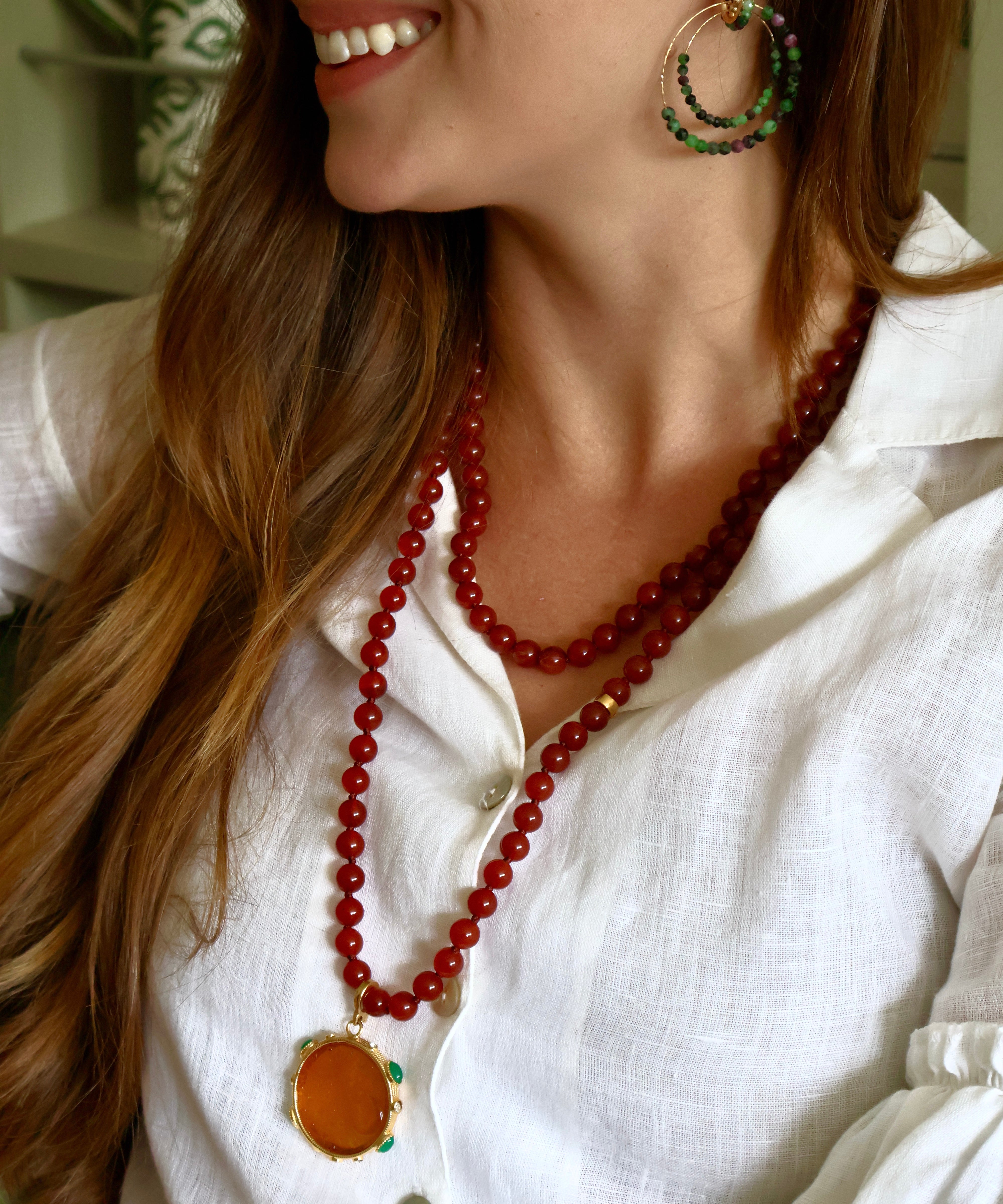 A close-up lifestyle shot of a woman wearing a long red beaded necklace with a large orange round pendant, styled with green and gold hoop earrings and a white linen shirt.