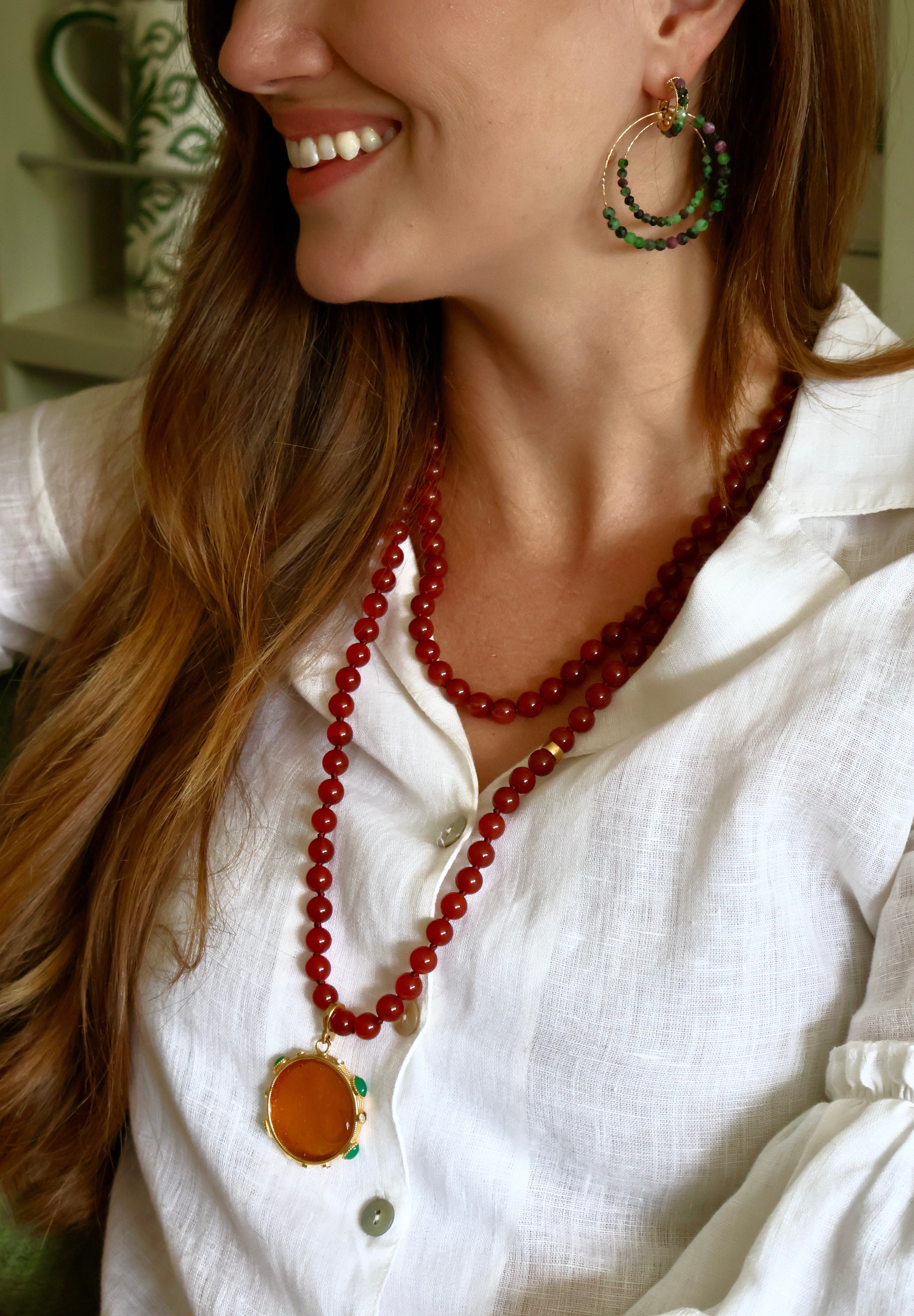 A close-up lifestyle shot of a woman wearing a long red beaded necklace with a large orange round pendant, styled with green and gold hoop earrings and a white linen shirt.