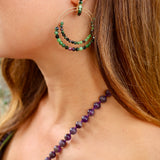 A close-up lifestyle shot of a woman wearing a delicate gold double-hoop earring adorned with green and dark beads, paired with a purple beaded necklace. A green strap is visible on her shoulder, adding a subtle pop of colour.