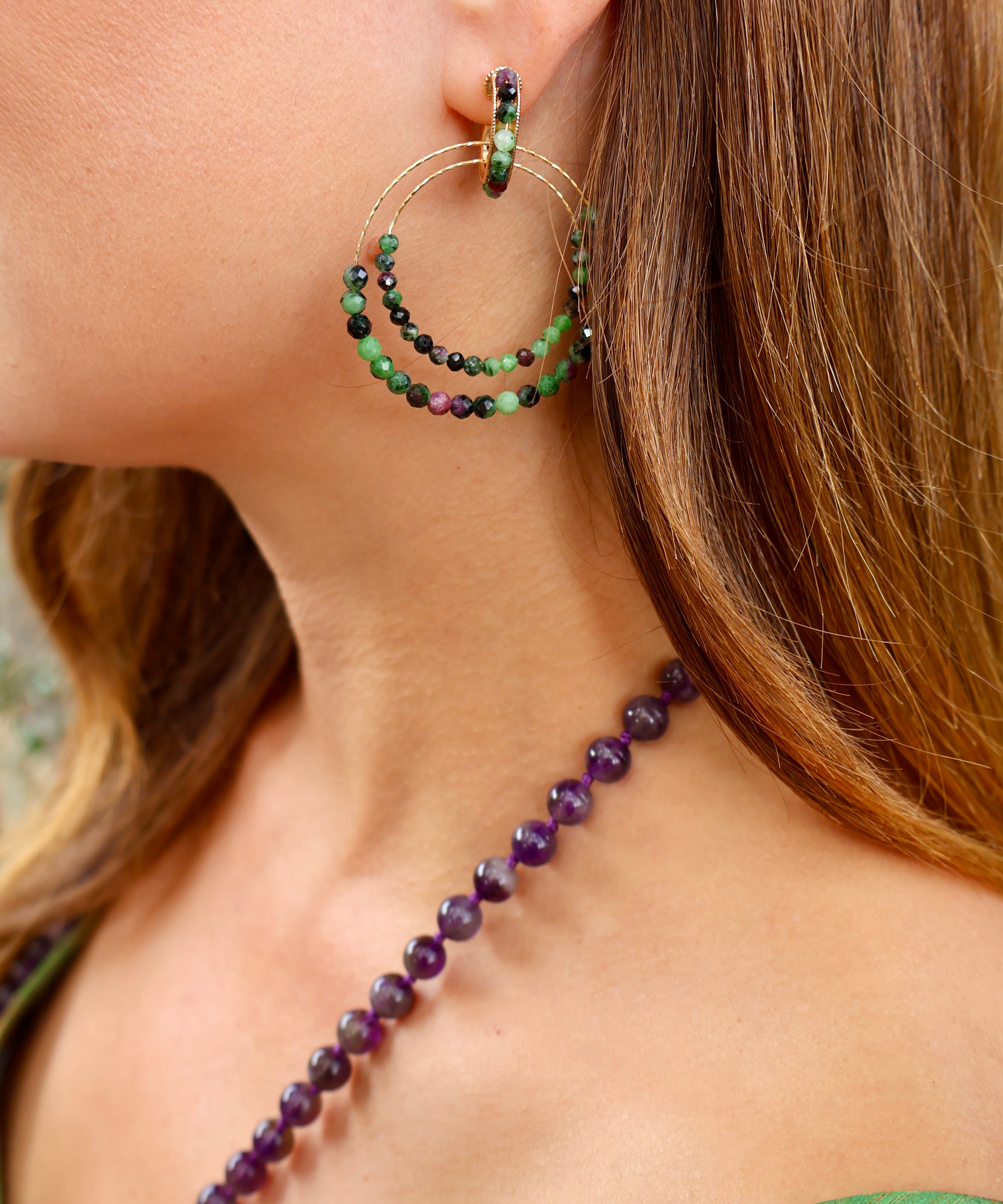 A close-up lifestyle shot of a woman wearing a delicate gold double-hoop earring adorned with green and dark beads, paired with a purple beaded necklace. A green strap is visible on her shoulder, adding a subtle pop of colour.