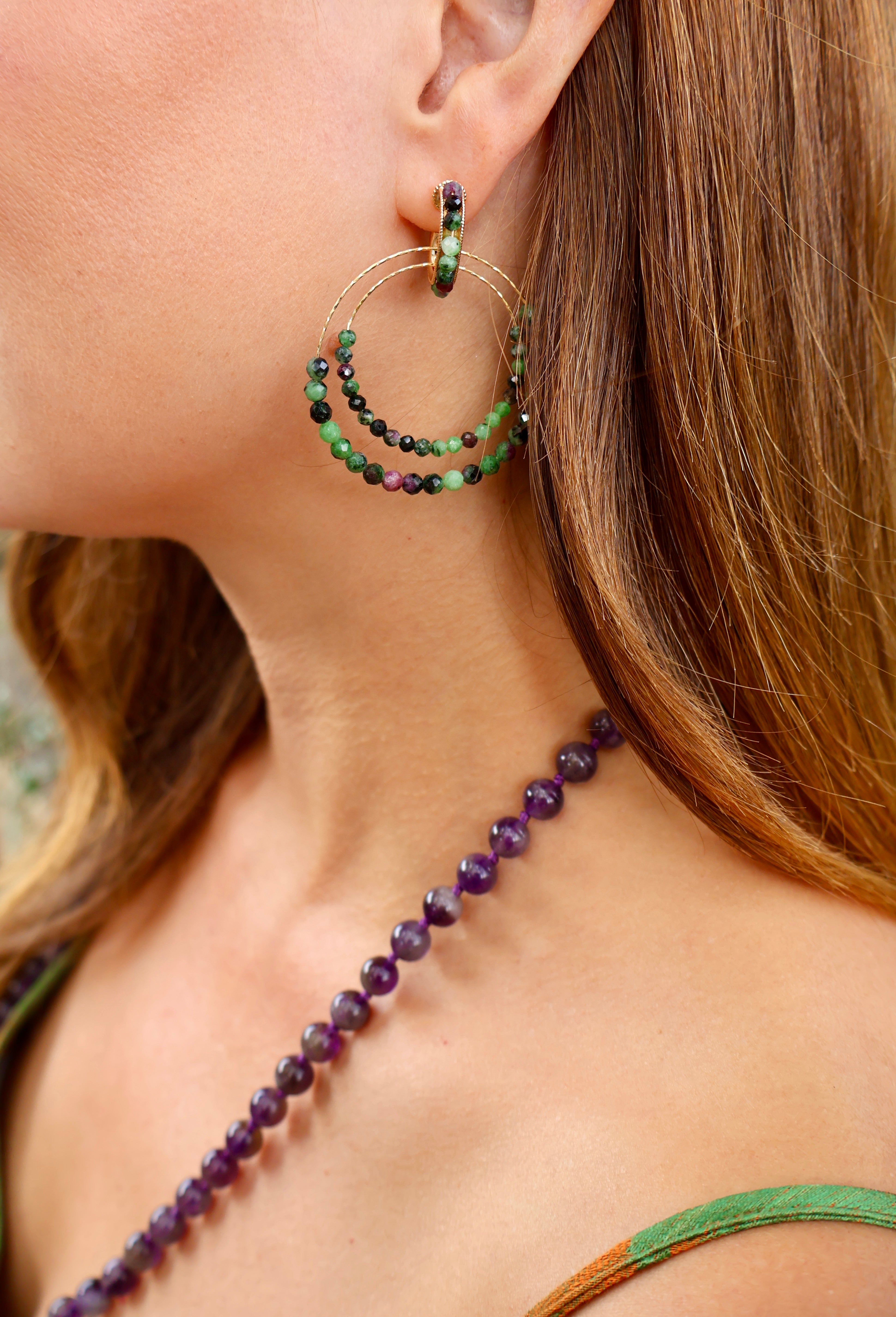 A close-up lifestyle shot of a woman wearing a delicate gold double-hoop earring adorned with green and dark beads, paired with a purple beaded necklace. A green strap is visible on her shoulder, adding a subtle pop of colour.