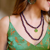 woman wearing a green strappy top, a purple beaded necklace with a small green charm attached and purple and green beaded statement hoop earrings. 