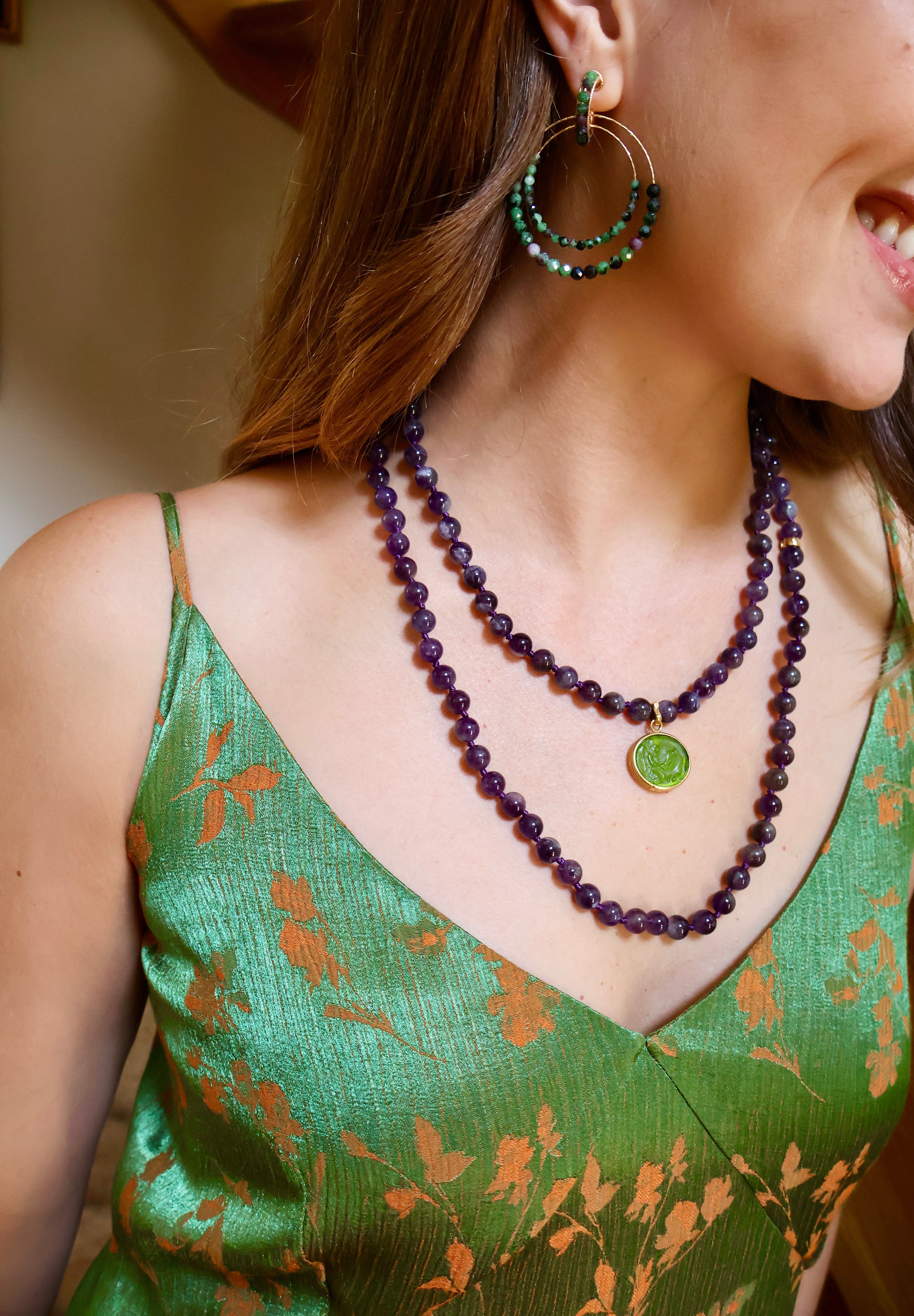 woman wearing a green strappy top, a purple beaded necklace with a small green charm attached and purple and green beaded statement hoop earrings. 