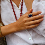 A close-up lifestyle shot of a person wearing a large oval ring with a deep blue stone and gold-toned setting, along with a red beaded necklace and a gold bracelet, all against a white linen shirt.