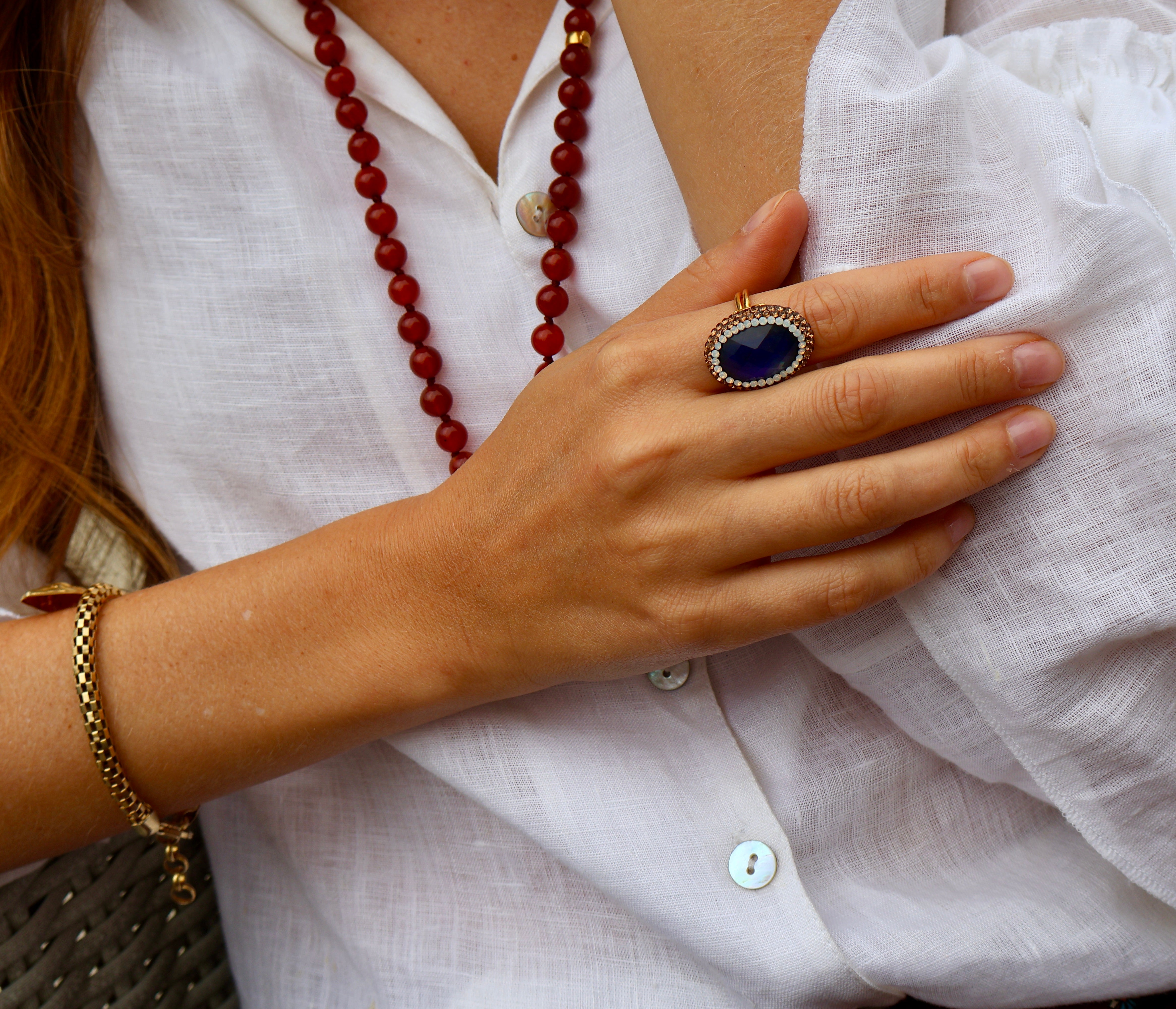 A close-up lifestyle shot of a person wearing a large oval ring with a deep blue stone and gold-toned setting, along with a red beaded necklace and a gold bracelet, all against a white linen shirt.