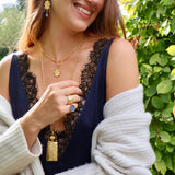 Photo of woman wearing gold coloured drop earrings and a matching necklace, a nother longer necklace with a gold coloured bottle charm and two rings. 