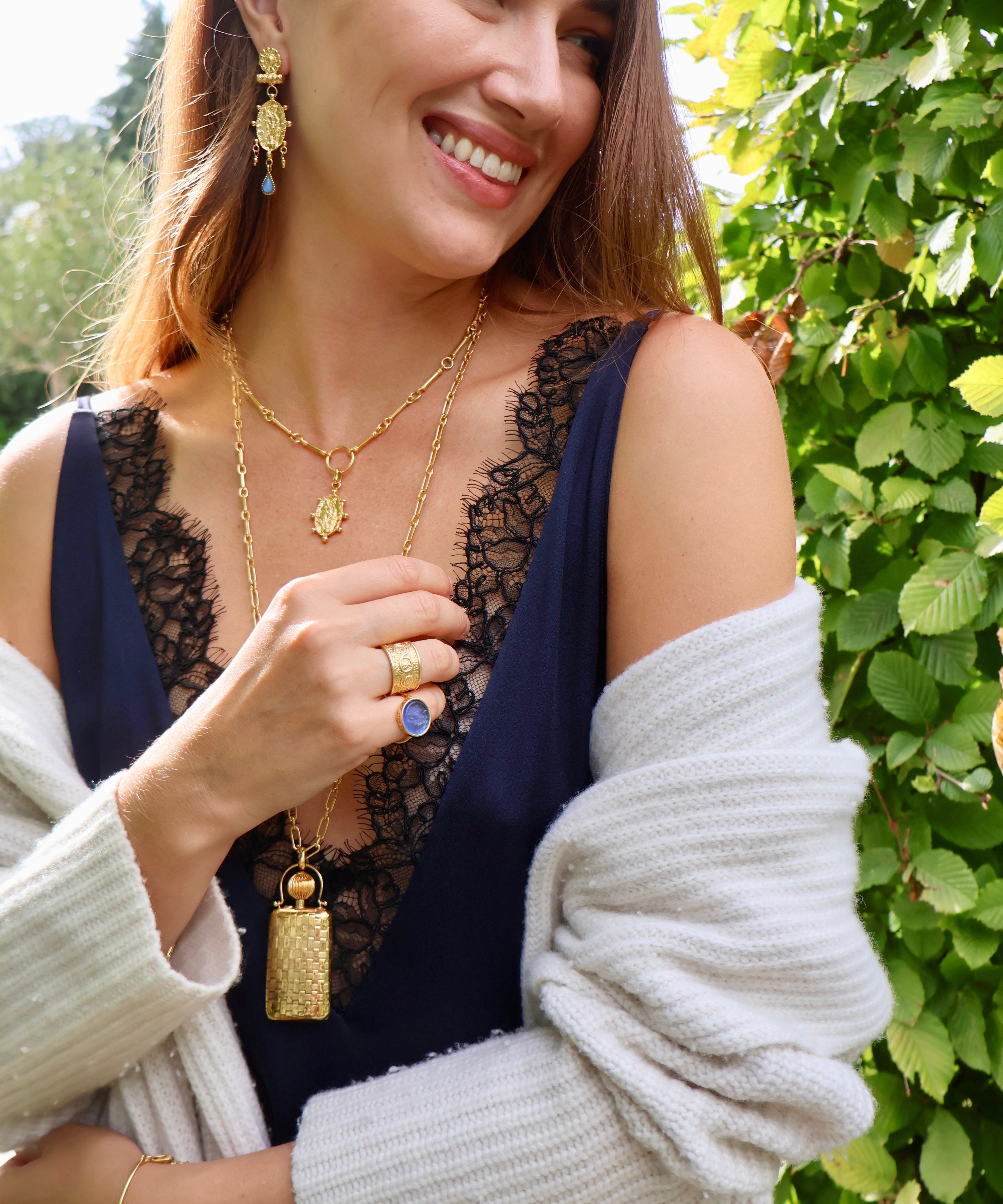 Photo of woman wearing gold coloured drop earrings and a matching necklace, a nother longer necklace with a gold coloured bottle charm and two rings. 