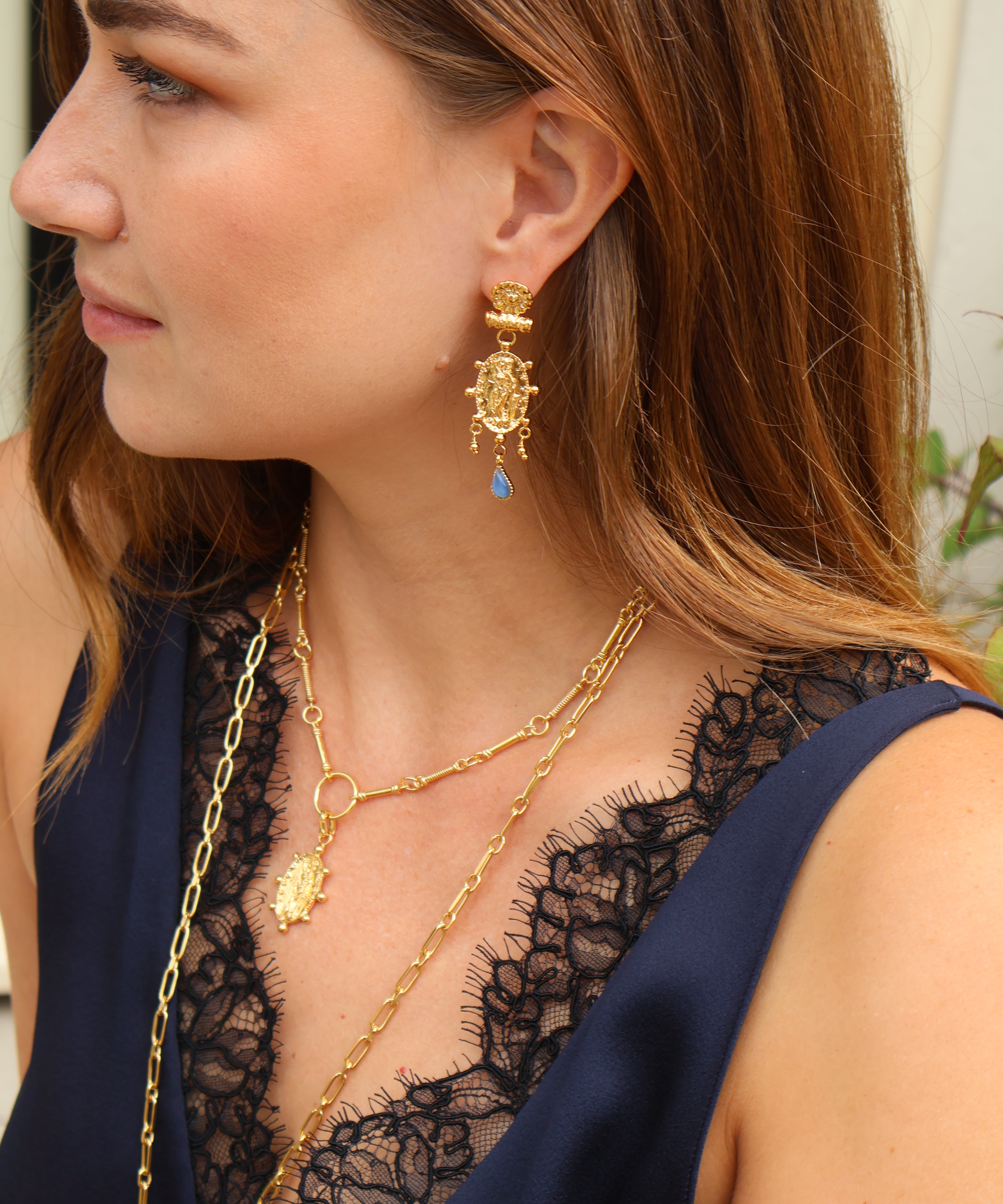 A close-up side profile of a woman wearing a gold dangle earring with a small blue stone, layered with gold necklaces, and wearing a navy blue top with black lace.