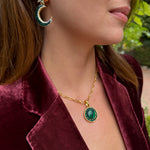 A close-up lifestyle shot of a woman wearing a gold chain necklace with a green oval pendant and a matching crescent moon and star earring, styled with a burgundy velvet blazer.