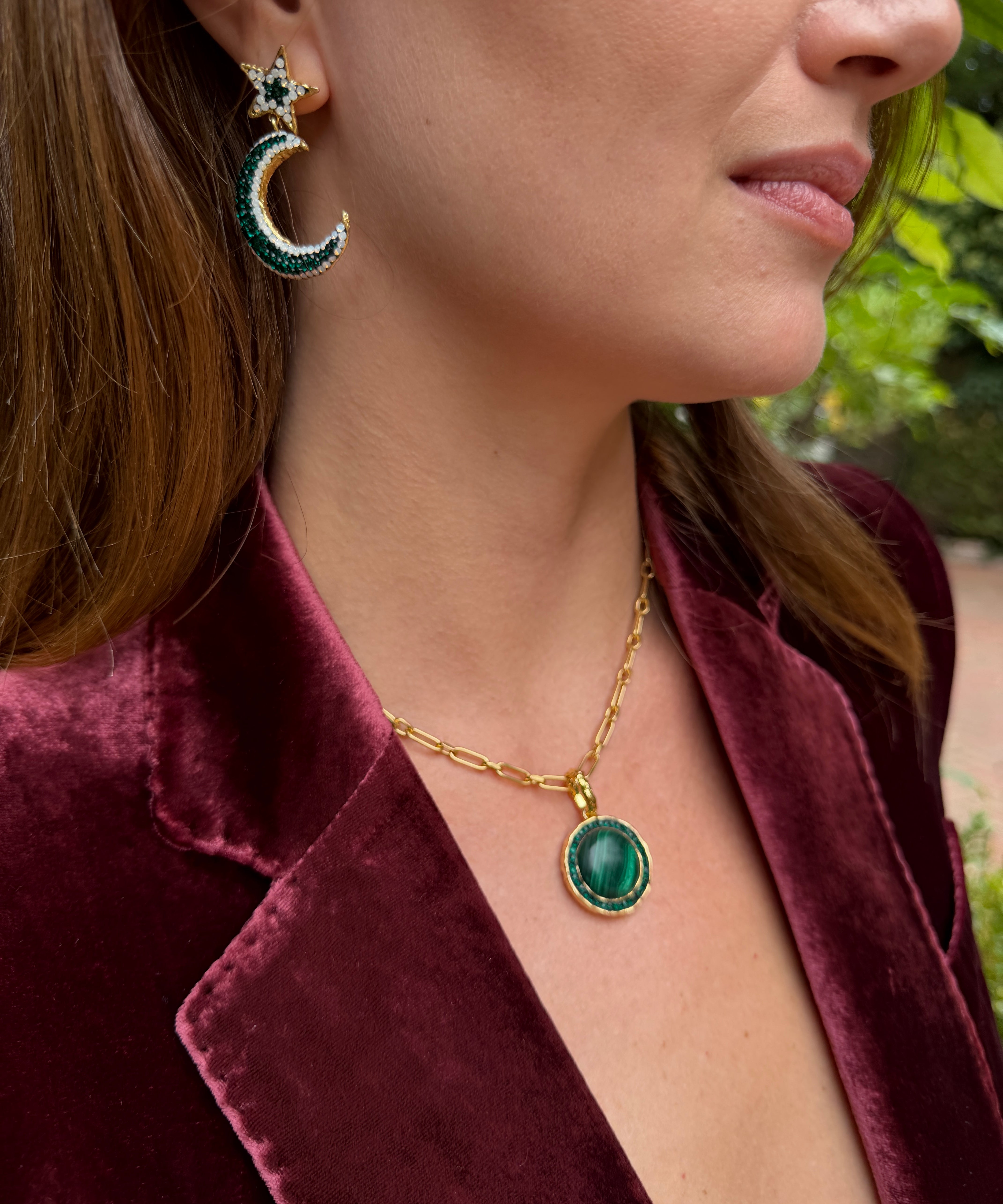 A close-up lifestyle shot of a woman wearing a gold chain necklace with a green oval pendant and a matching crescent moon and star earring, styled with a burgundy velvet blazer.