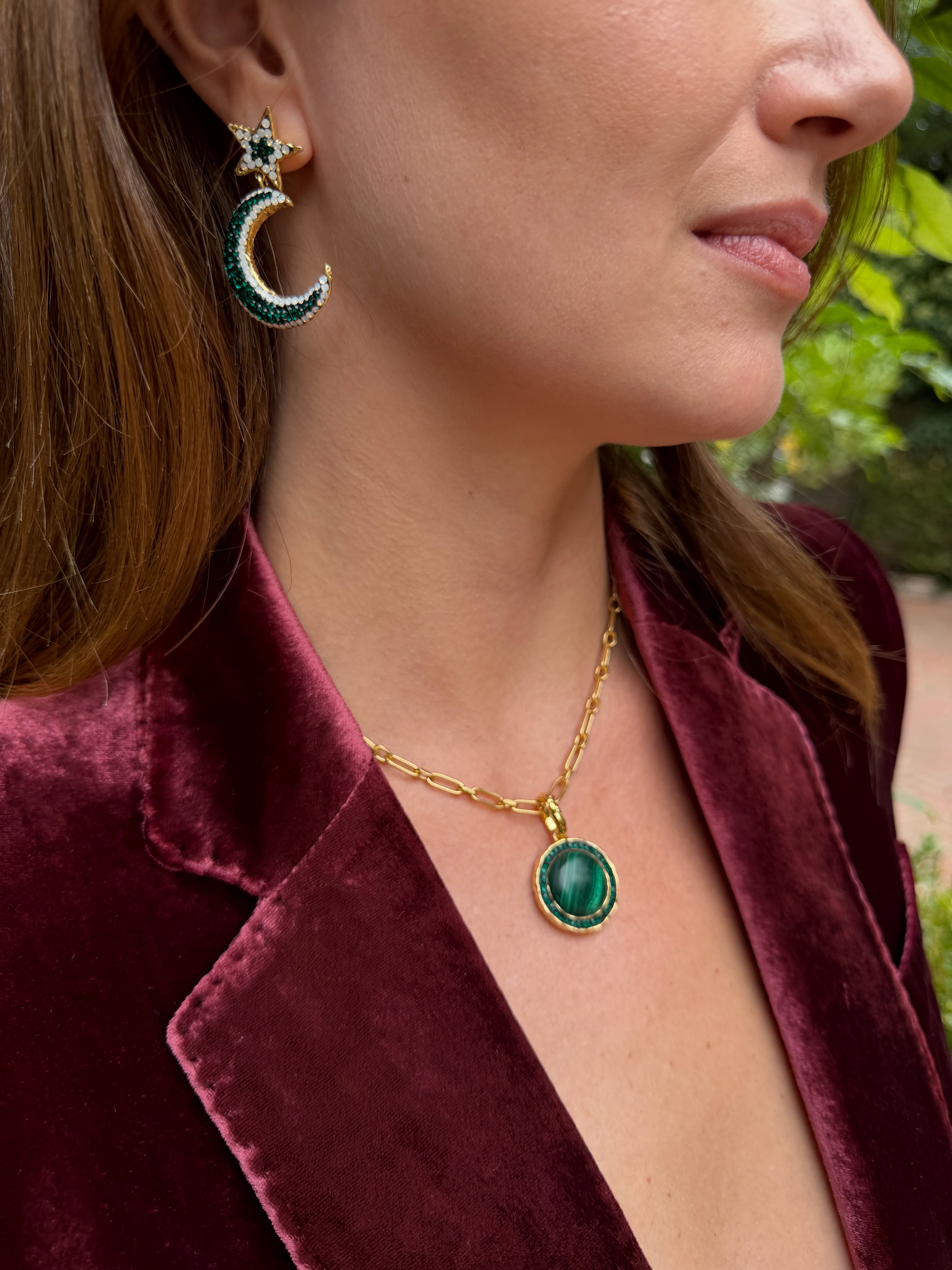 A close-up lifestyle shot of a woman wearing a gold chain necklace with a green oval pendant and a matching crescent moon and star earring, styled with a burgundy velvet blazer.