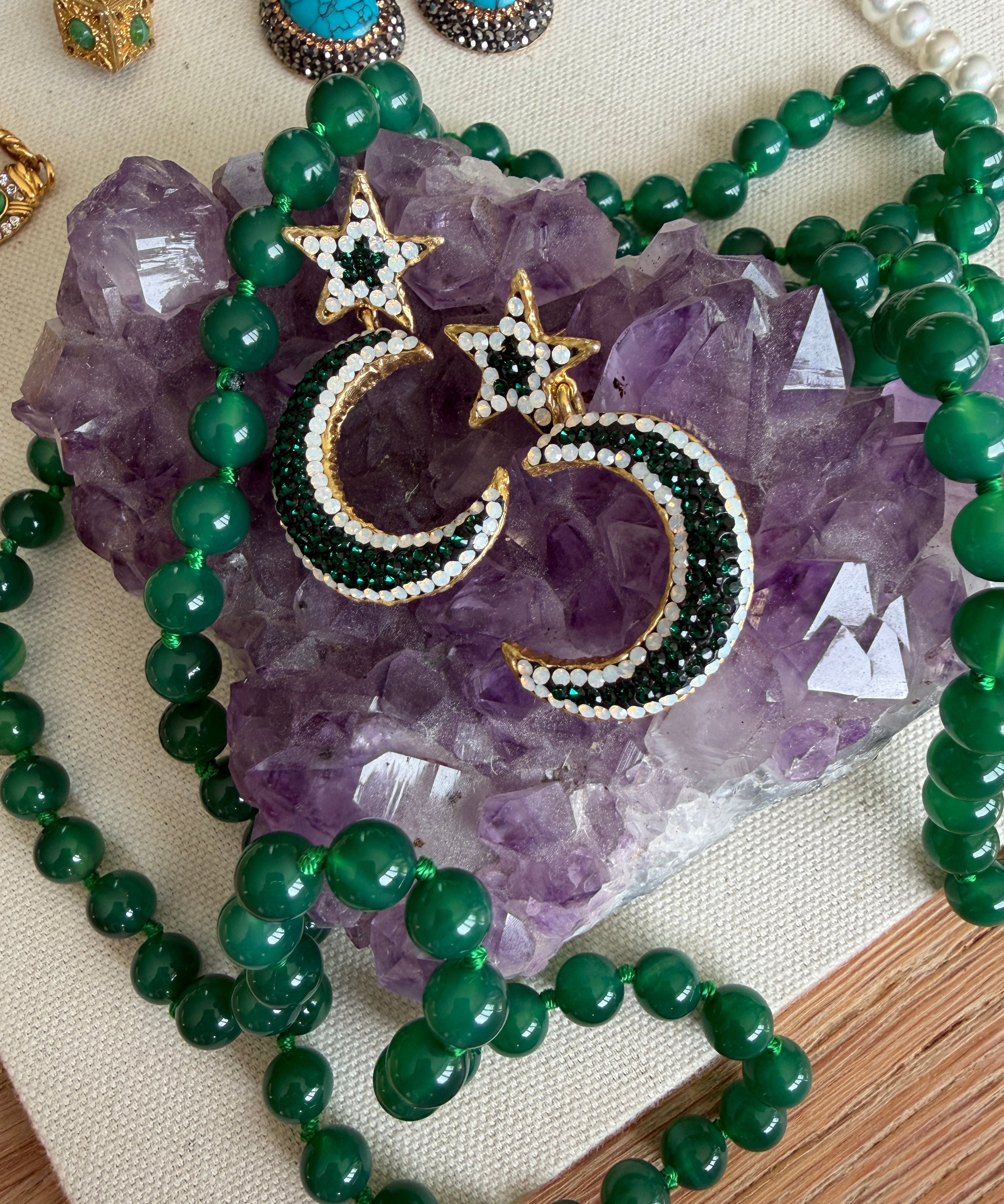 Gold crescent moon and star earrings embellished with green and white beads, displayed on a purple amethyst crystal cluster with a green beaded necklace.