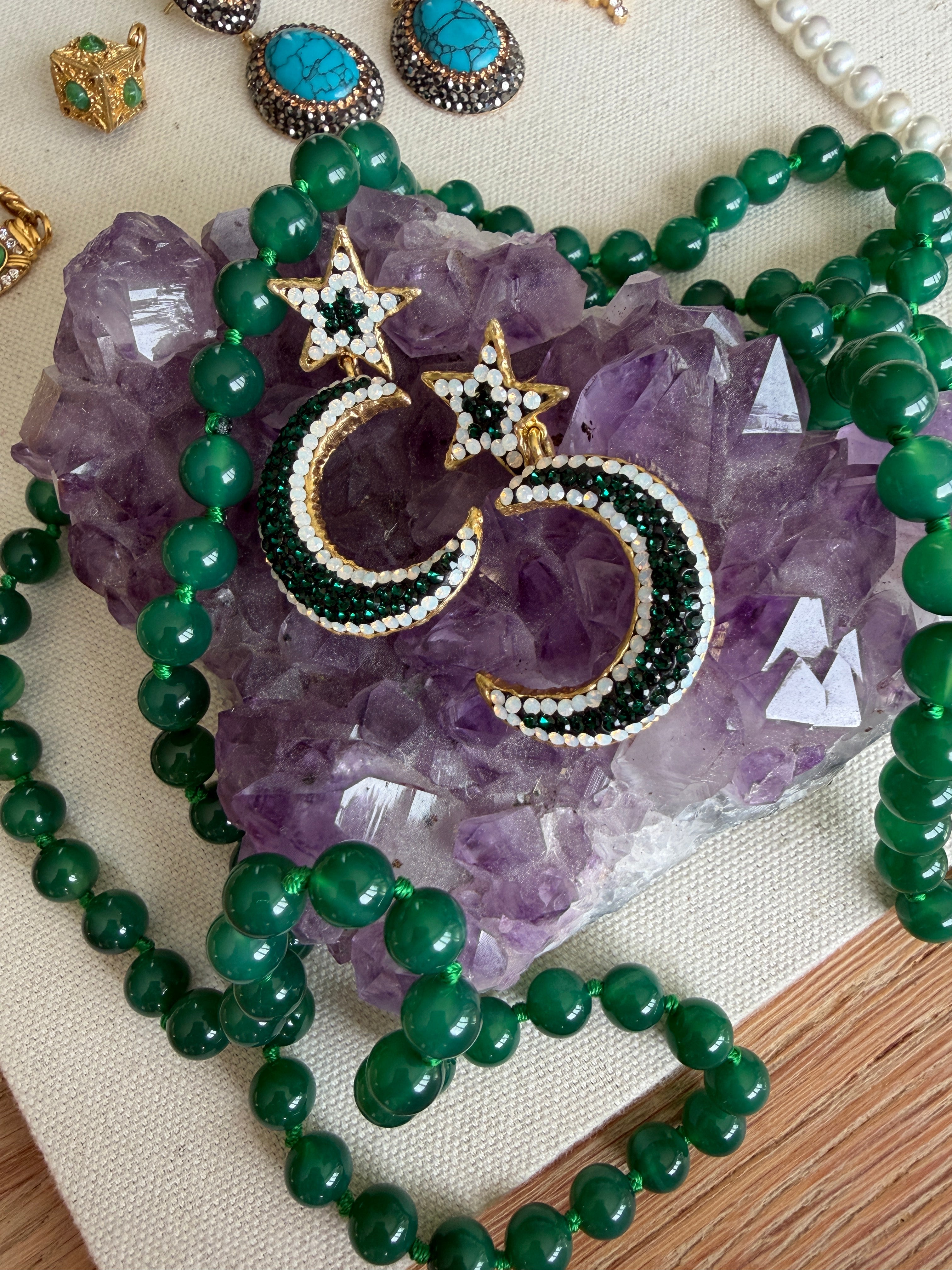 Gold crescent moon and star earrings embellished with green and white beads, displayed on a purple amethyst crystal cluster with a green beaded necklace.