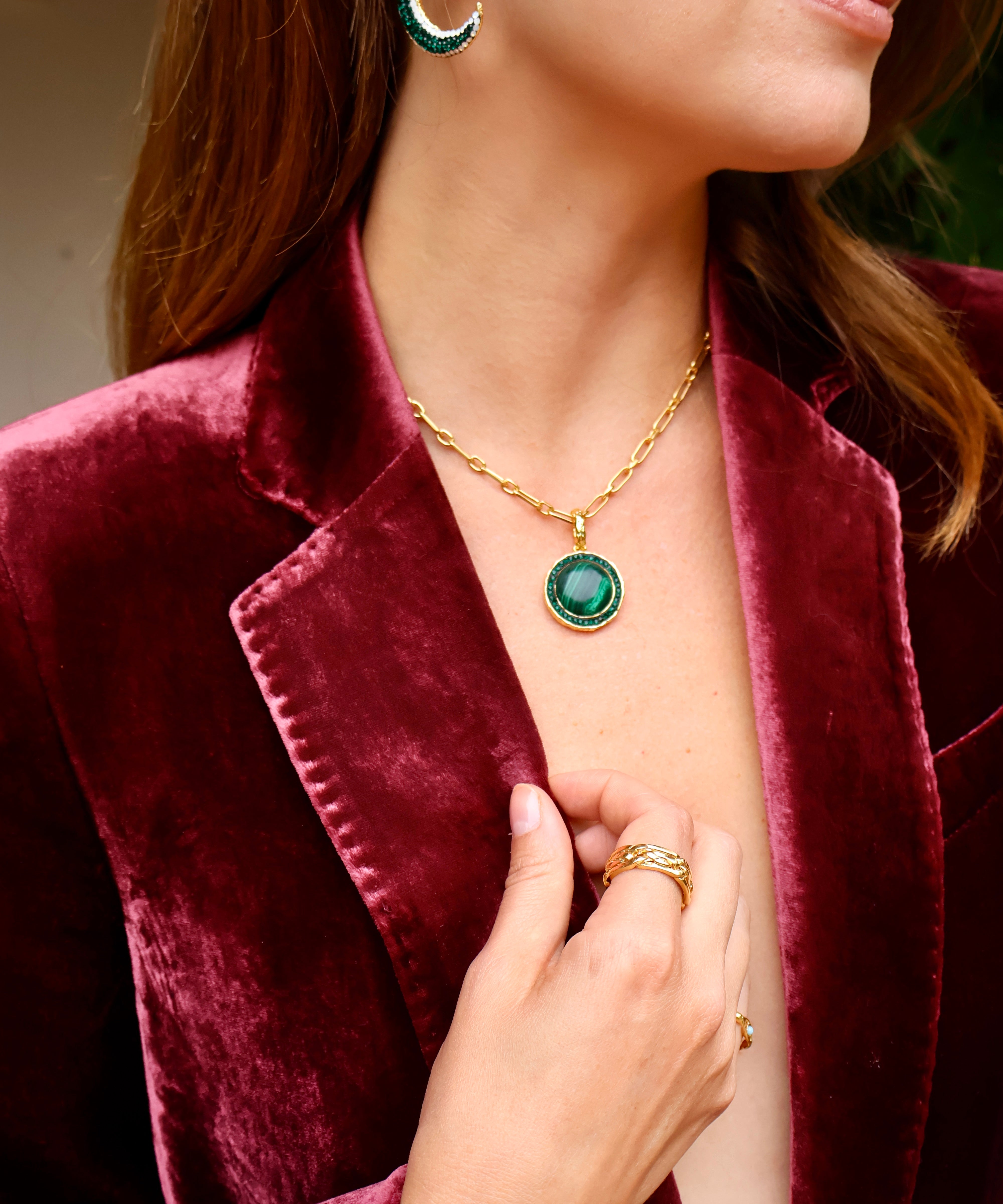 A close-up lifestyle shot of a woman wearing a gold chain necklace with a round green gemstone pendant, styled with a burgundy velvet blazer.