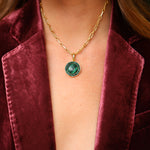 A gold chain necklace featuring a smooth, round green malachite pendant surrounded by sparkling crystals, styled beautifully with a luxurious burgundy velvet blazer.
