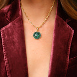 A gold chain necklace featuring a smooth, round green malachite pendant surrounded by sparkling crystals, styled beautifully with a luxurious burgundy velvet blazer.